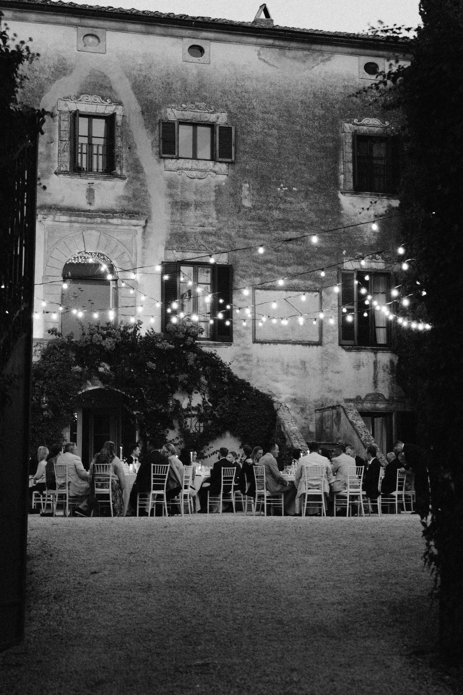 String lights illuminate an outdoor reception area with white chairs against a historic building.