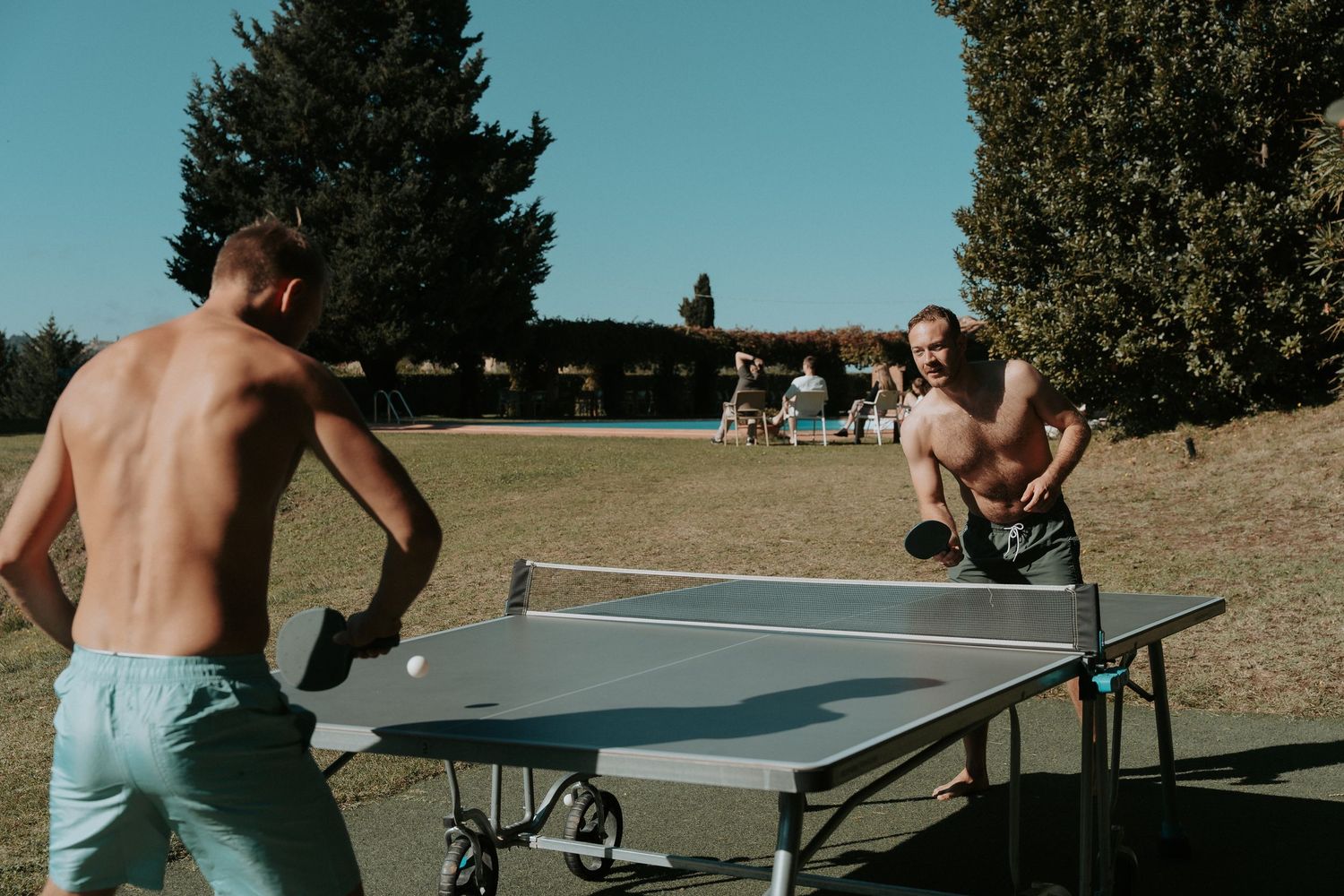 People playing ping pong outdoors on multiple tables with trees in background.