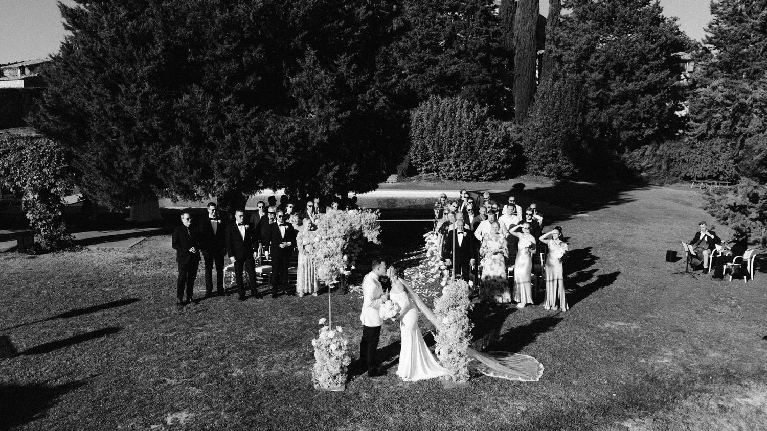 A wedding ceremony takes place outdoors on a sunny day with guests seated in white chairs.