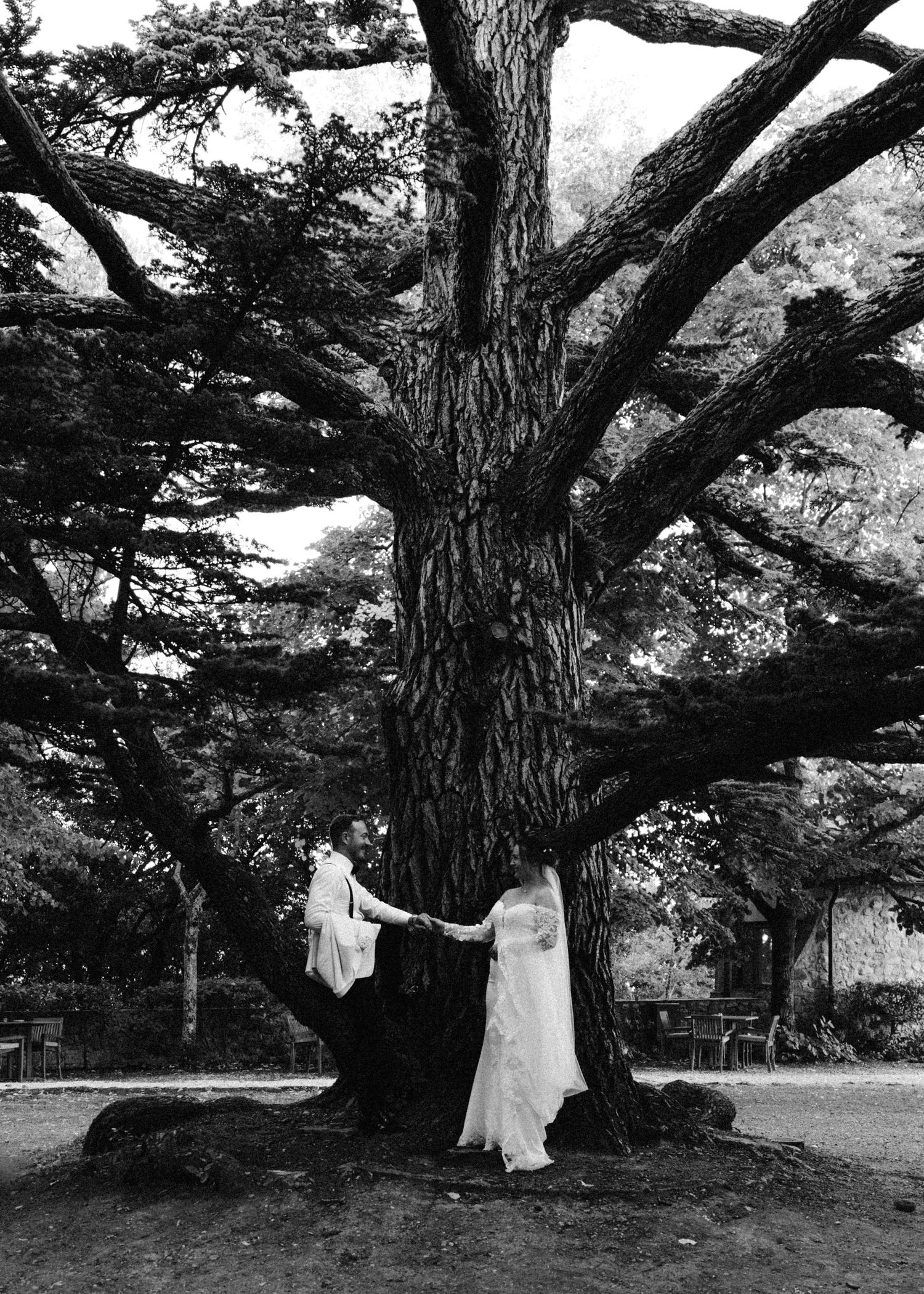Two figures in white dresses dance around a large tree trunk in a black and white photograph.