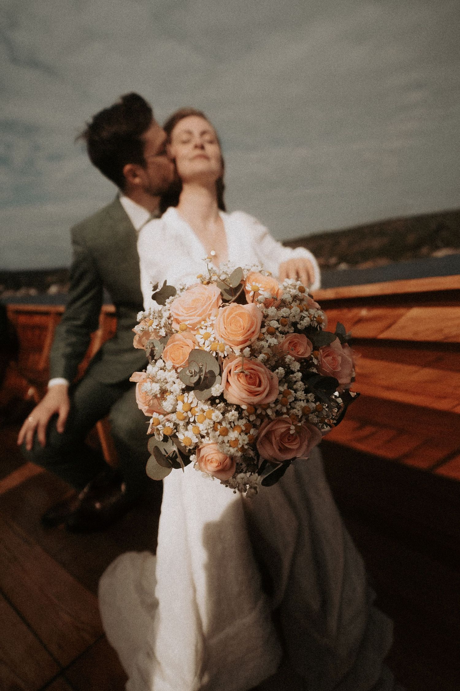 A romantic wedding couple embraces on a deck while holding a peach and blush floral bouquet at sunset.