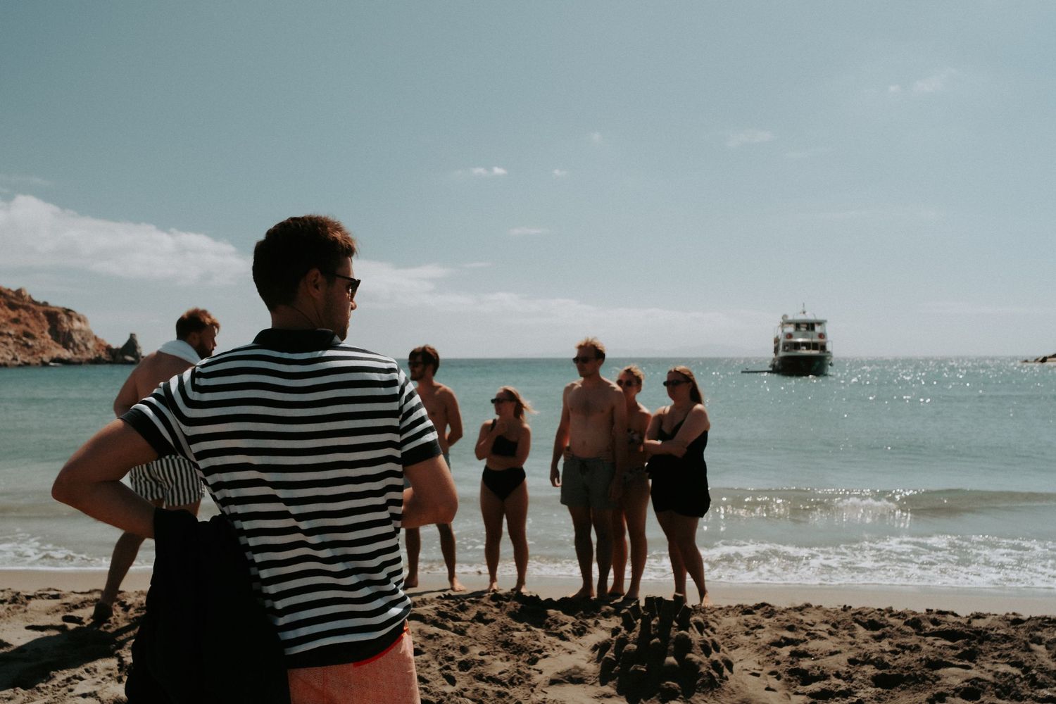 Beachgoers gather on a rocky shore with a boat visible in the distance.