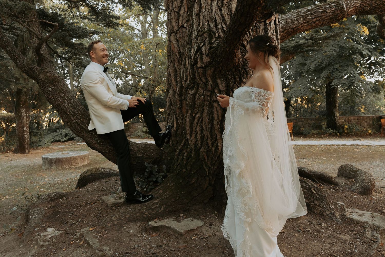 Wedding couple poses playfully by a large tree trunk in a natural setting.