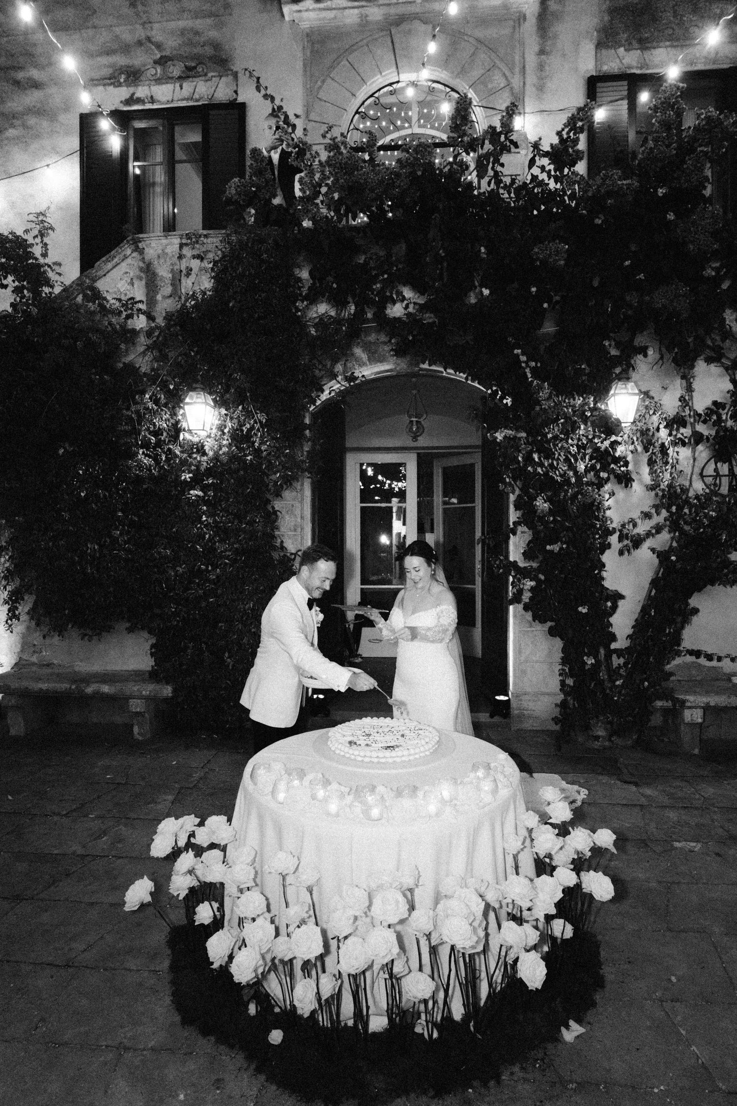Elegant wedding cake cutting scene at night under an arched doorway with twinkling lights.