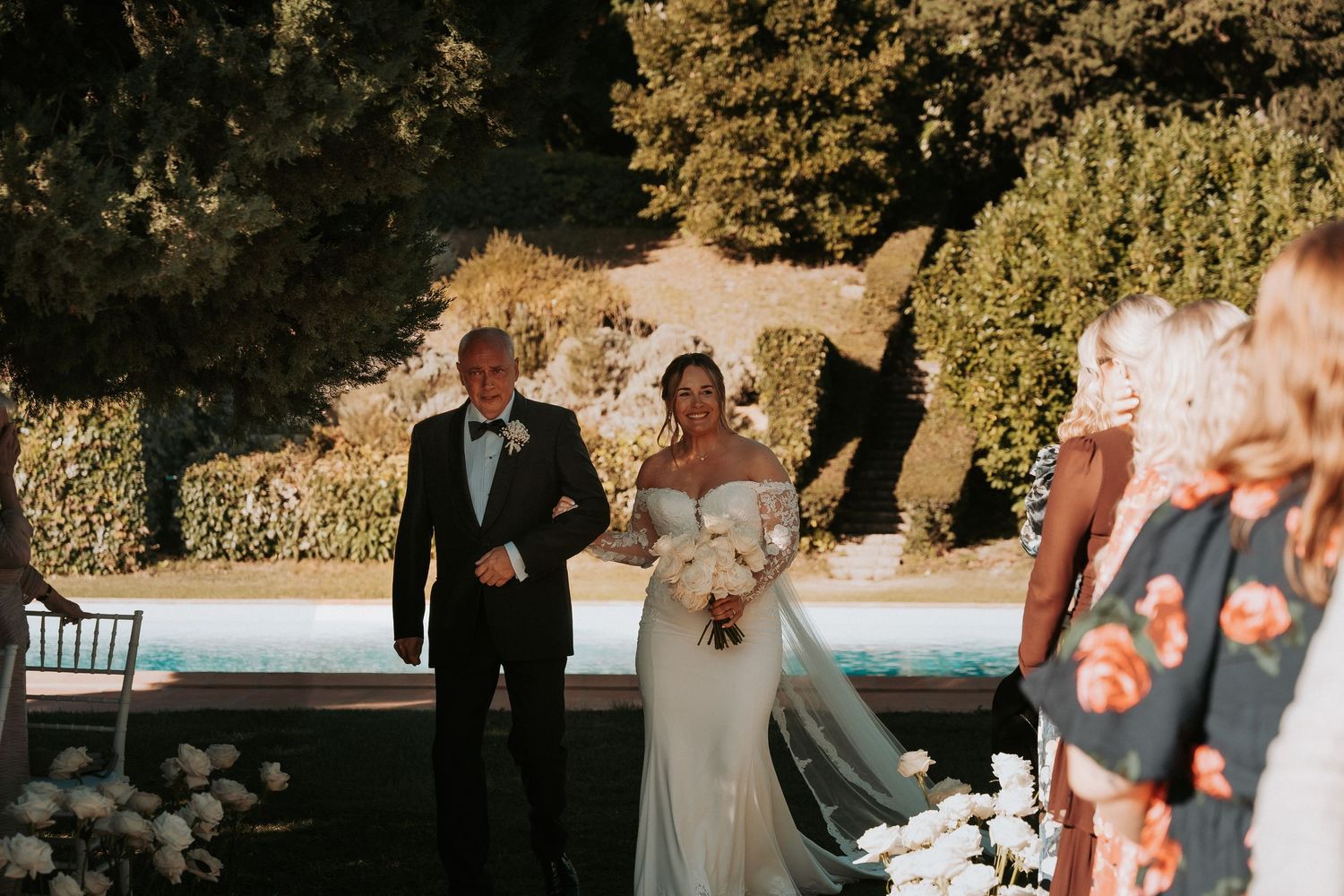 A wedding ceremony takes place outdoors with formal attire and floral decorations on a sunny day.
