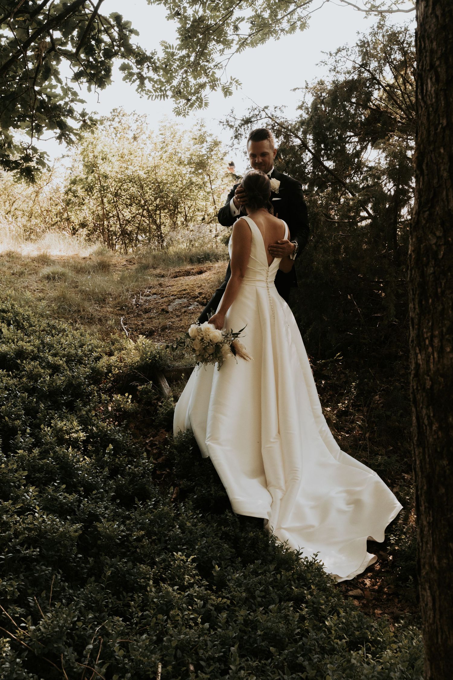 A romantic wedding couple embraces in a sun-dappled forest setting wearing elegant white attire.