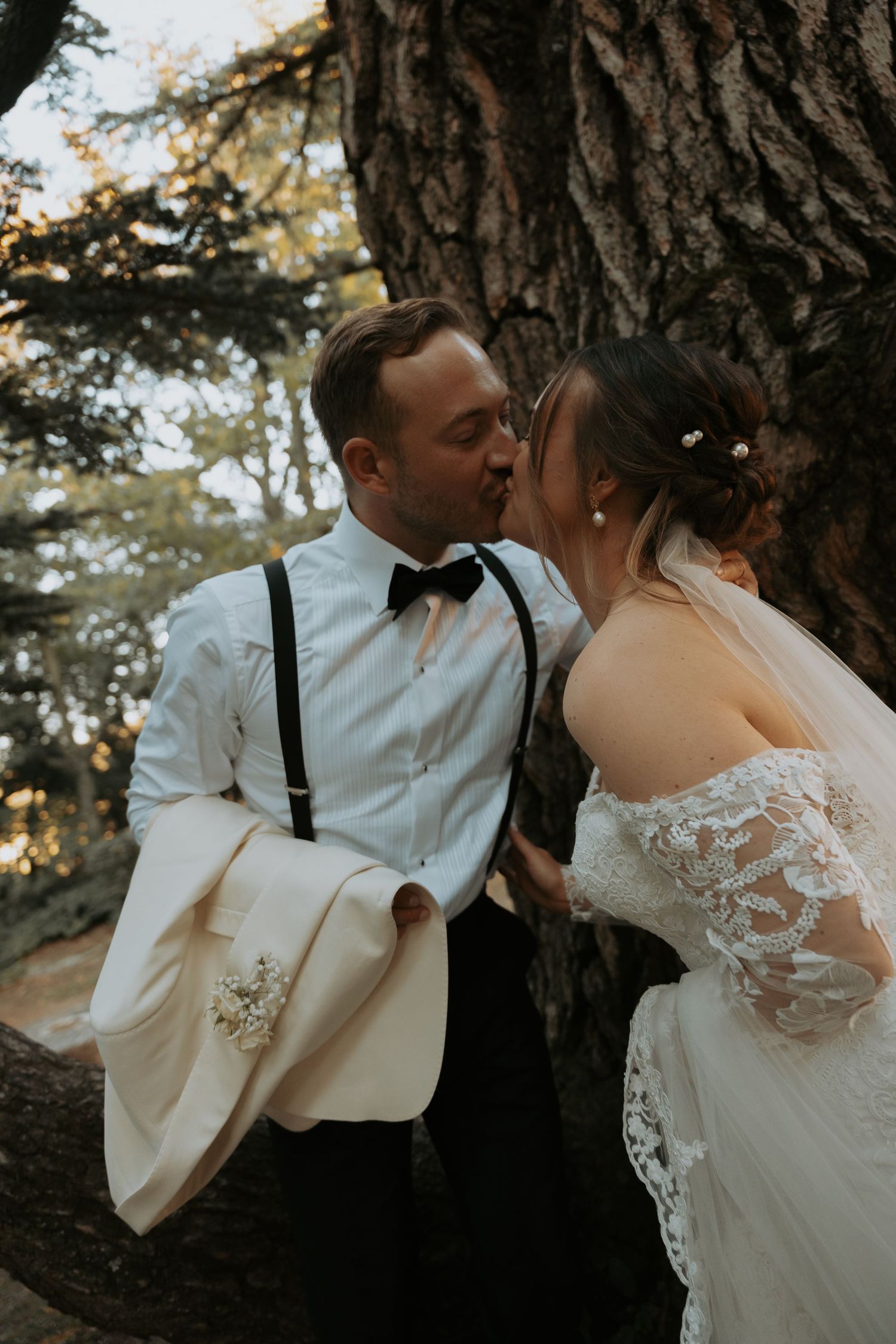 A tender moment between newlyweds in formal attire against a rustic tree backdrop.