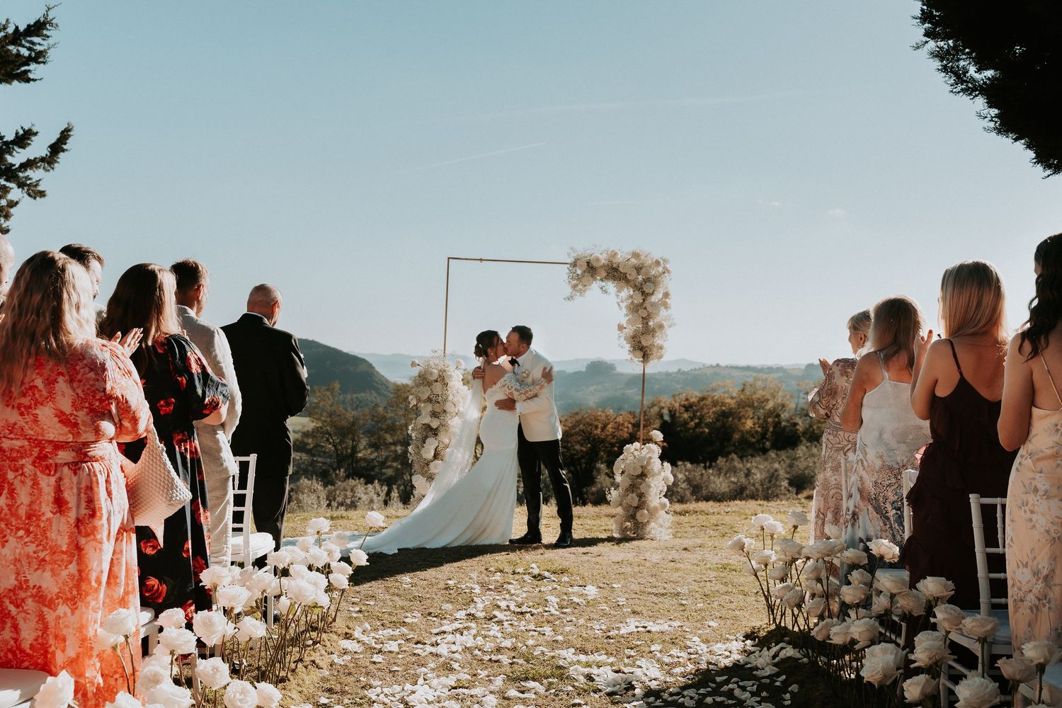 Wedding ceremony with guests standing along flower-lined aisle under clear blue sky.