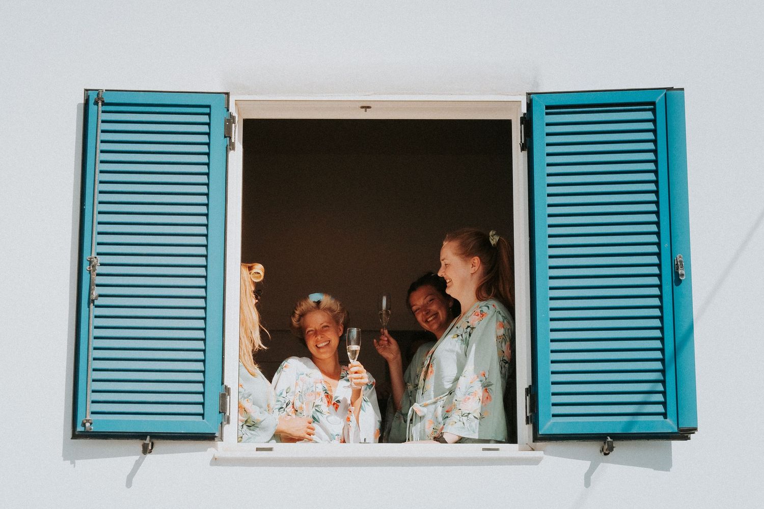 Bridal party in robes looking out of a window with turquoise shutters.