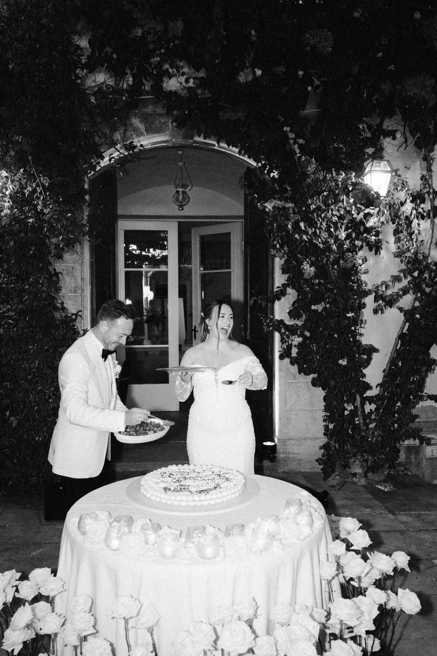 Black and white photo of a couple enjoying their wedding cake under an elegant archway.