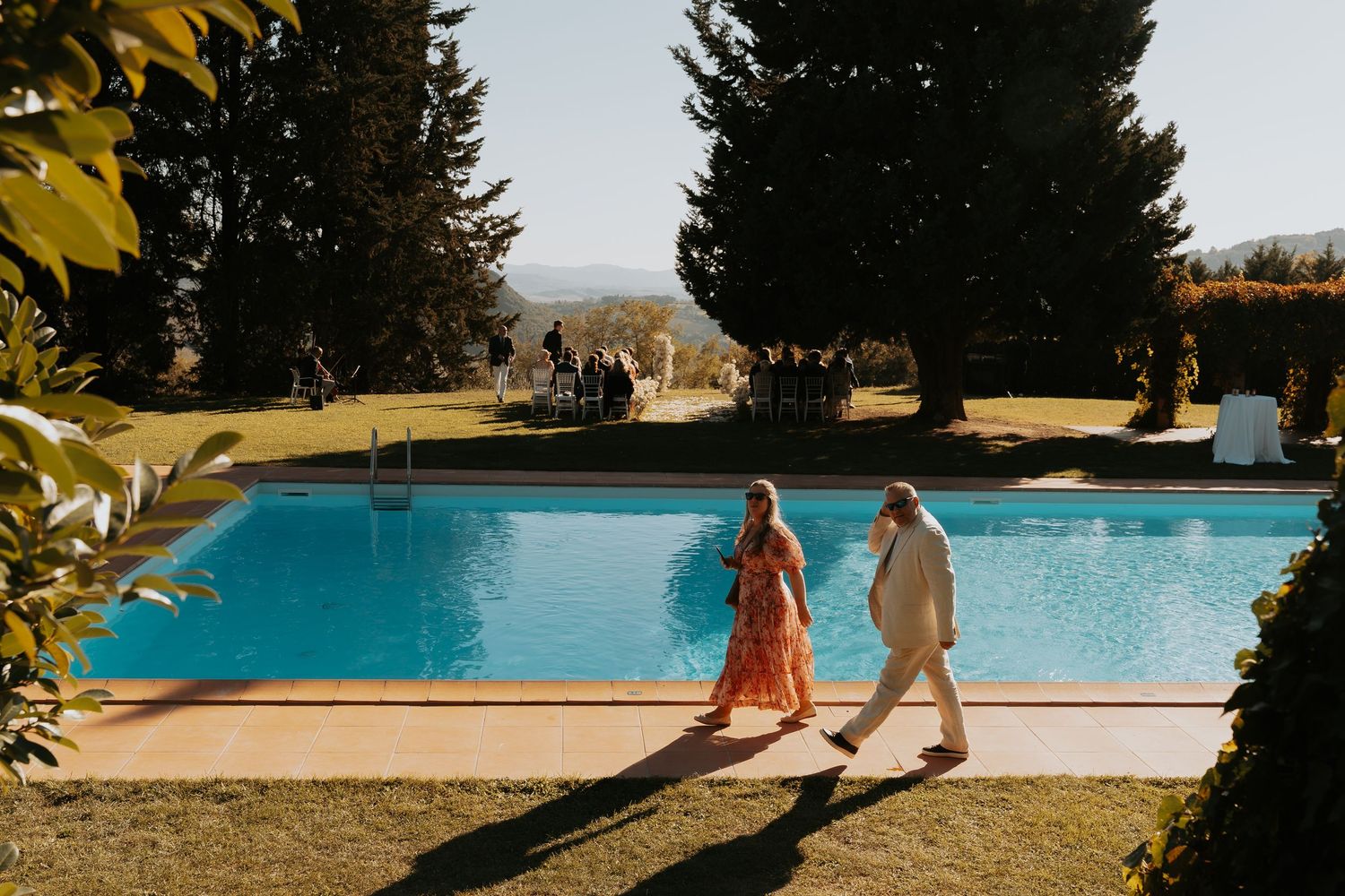 People walking along the edge of a swimming pool surrounded by pine trees at sunset.