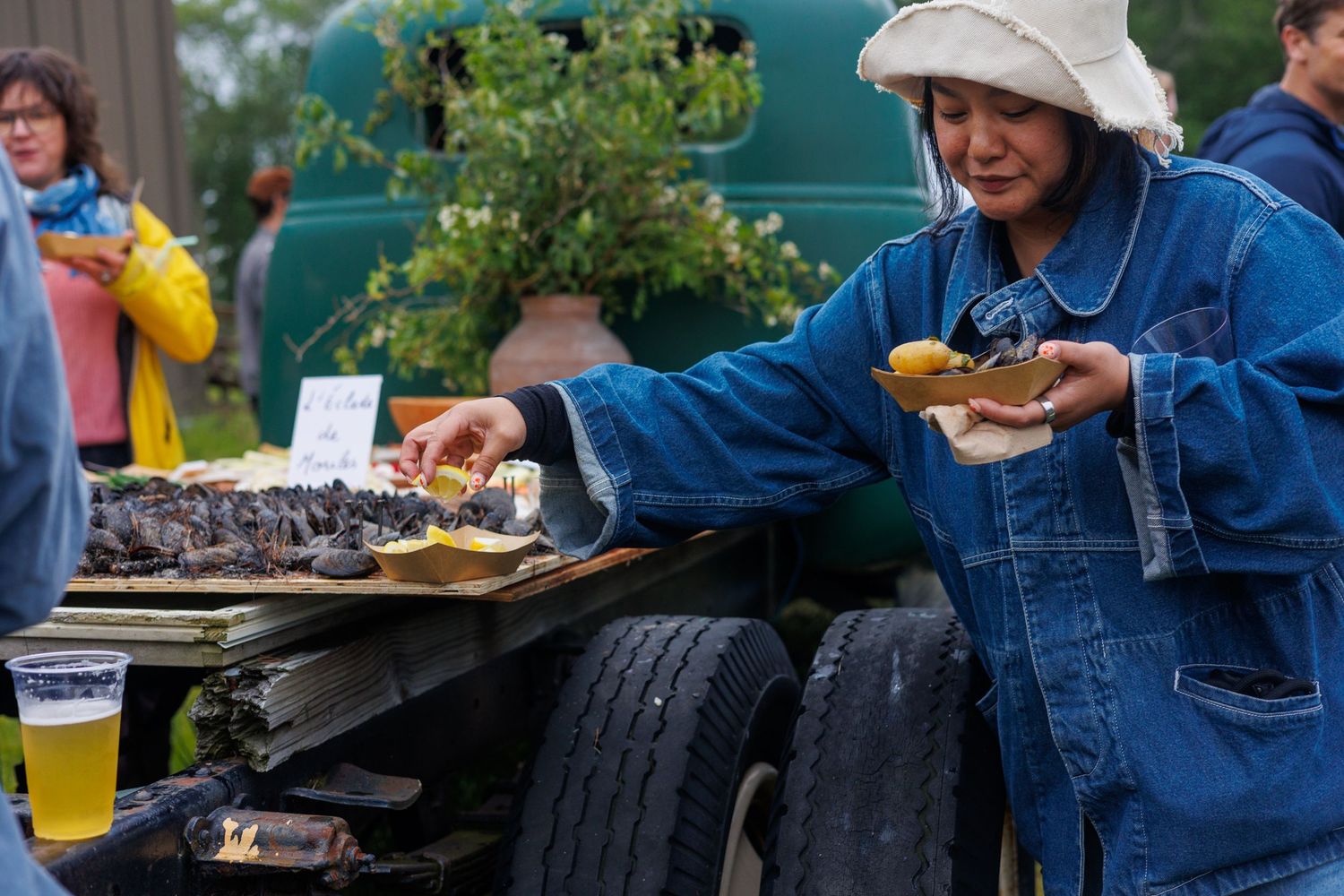 Someone in denim coveralls shucks fresh oysters at an outdoor event with a makeshift table made from tires. north shore massachusetts