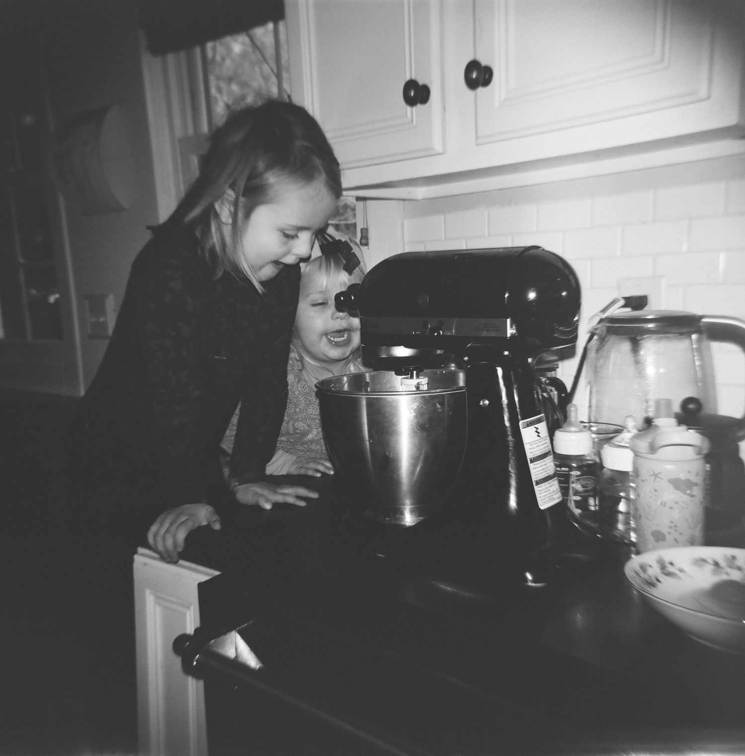 two children leans over a stand mixer on a kitchen counter in a black and white photograph. north shore massachusetts