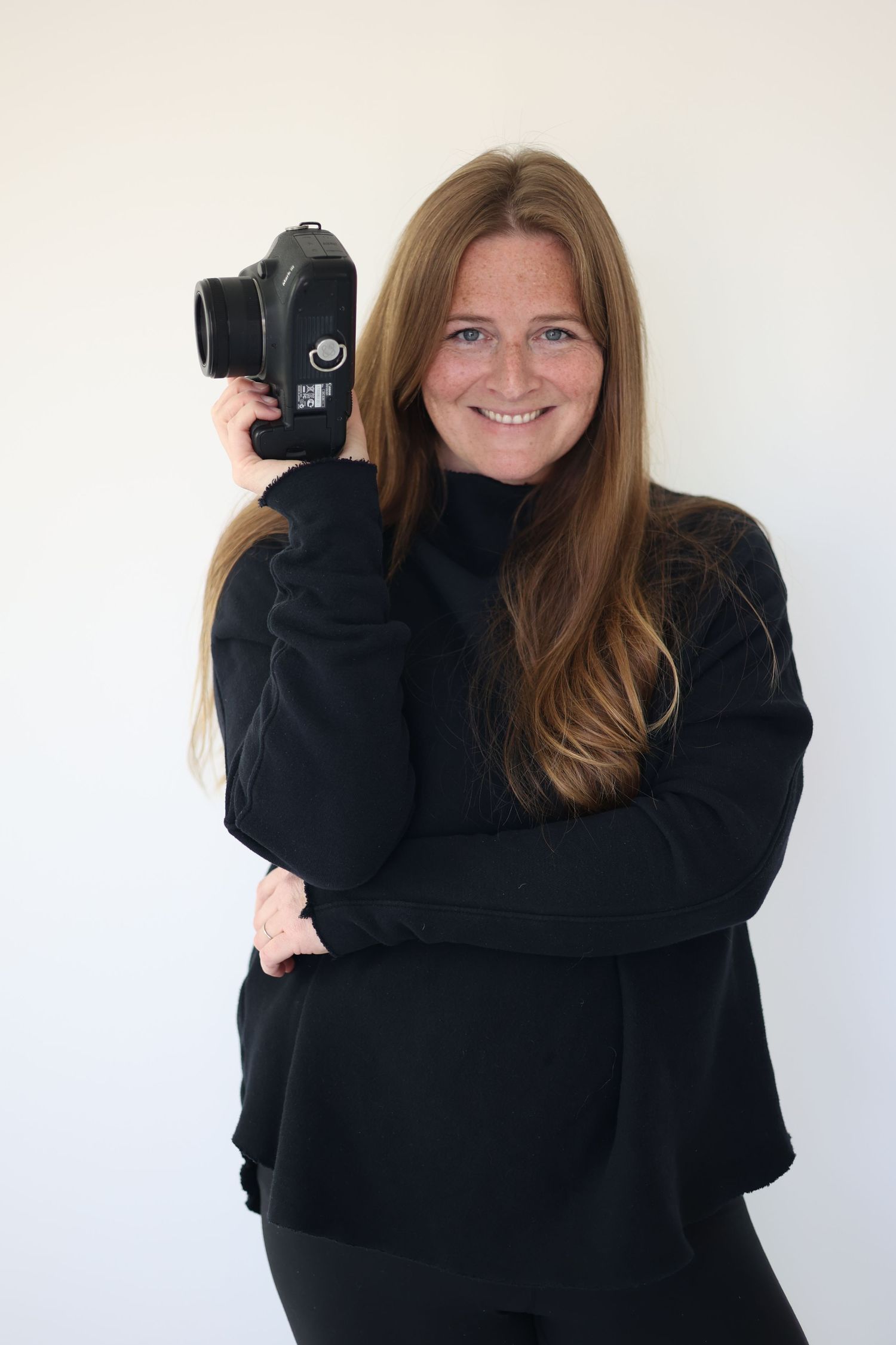 a photographer in a black sweater holding a DSLR camera against a white background. north shore massachusetts