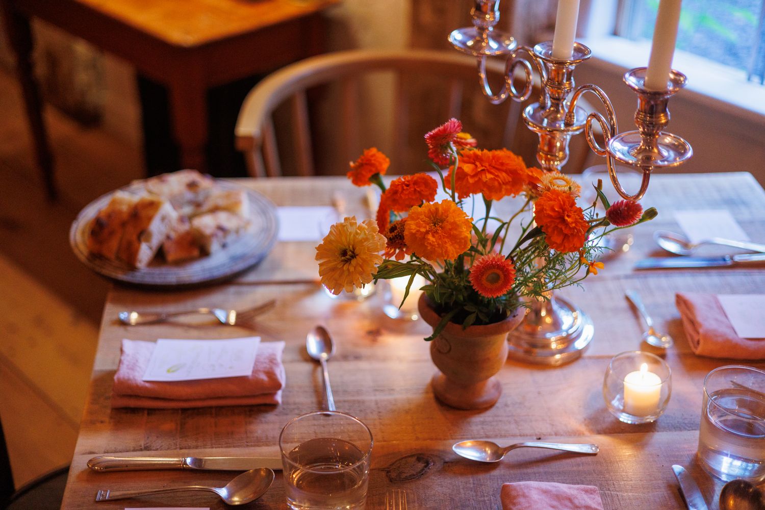 A cozy dining table set with orange flowers in a clay vase, candles, and place settings for an intimate gathering. north shore massachusetts