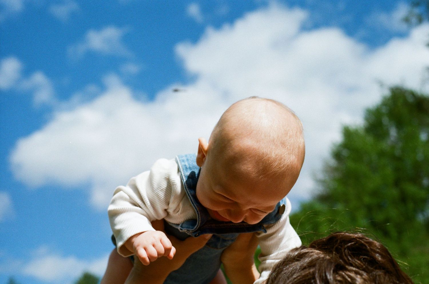 A baby in a white sweater is lifted up against a bright blue sky with fluffy white clouds. north shore massachusetts