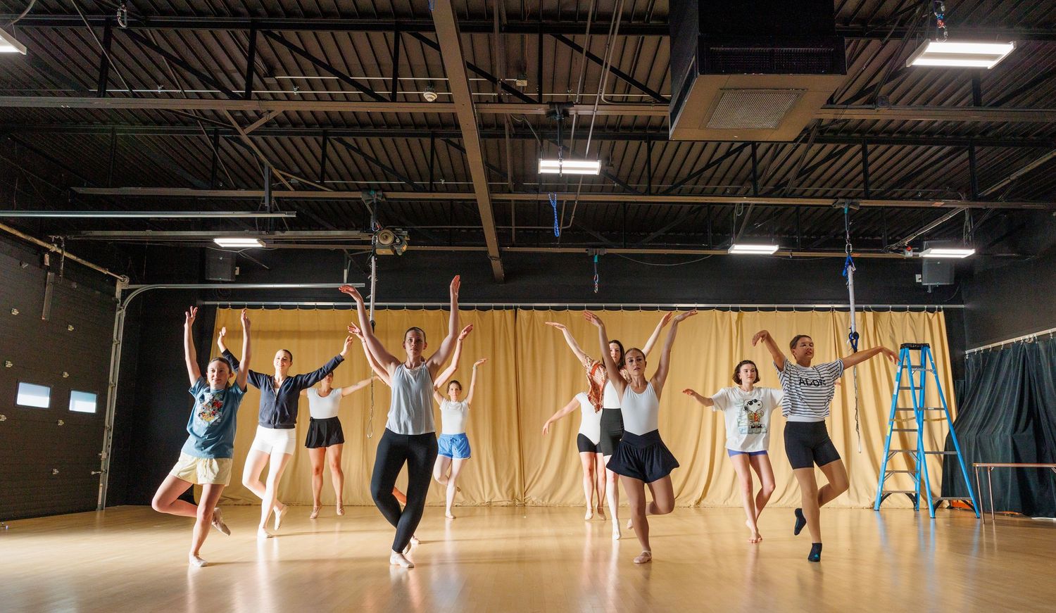 Dancers leap joyfully in unison across a wooden floor studio with high ceilings and stage curtains. ipswich massachusetts