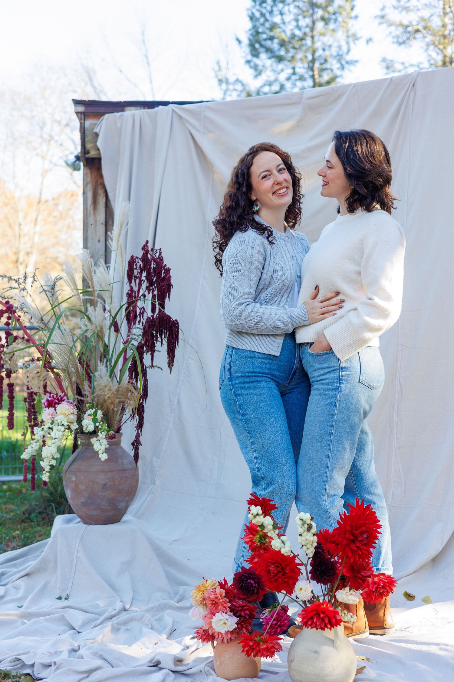Two people in sweaters and jeans share a joyful moment against a white backdrop with colorful floral arrangements. north shore massachusetts