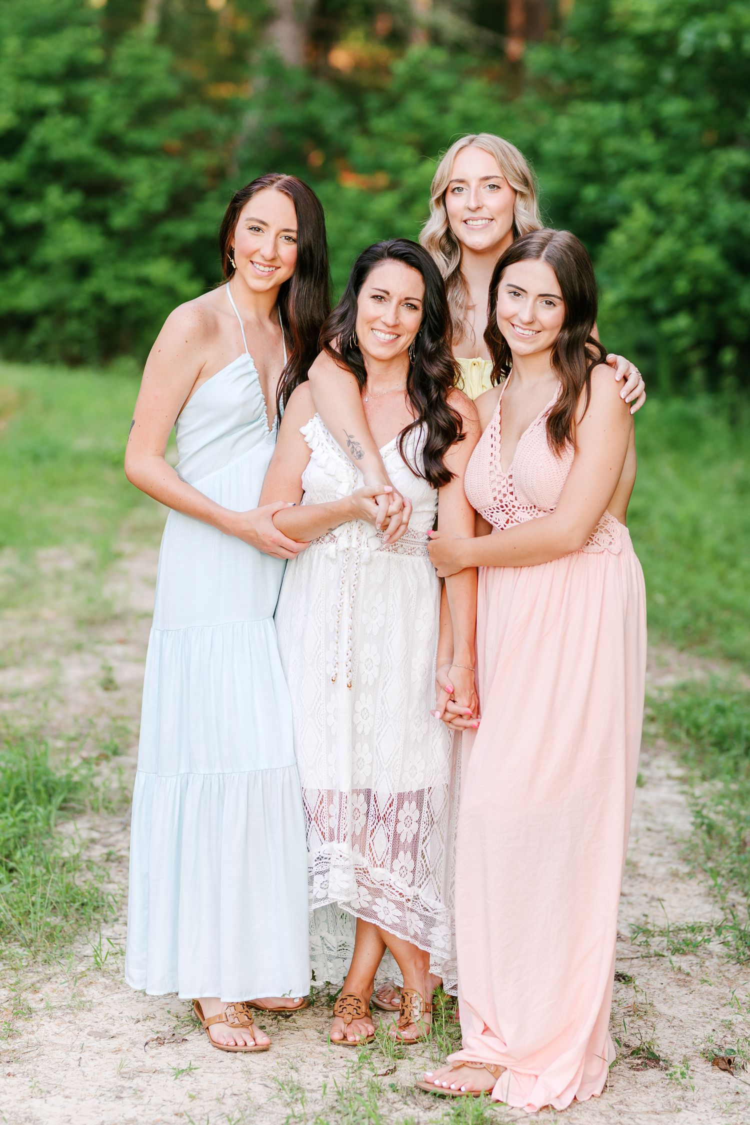 Three women in pastel dresses smile and embrace while standing in a lush green outdoor setting in Winston Salem with a Family Photographer