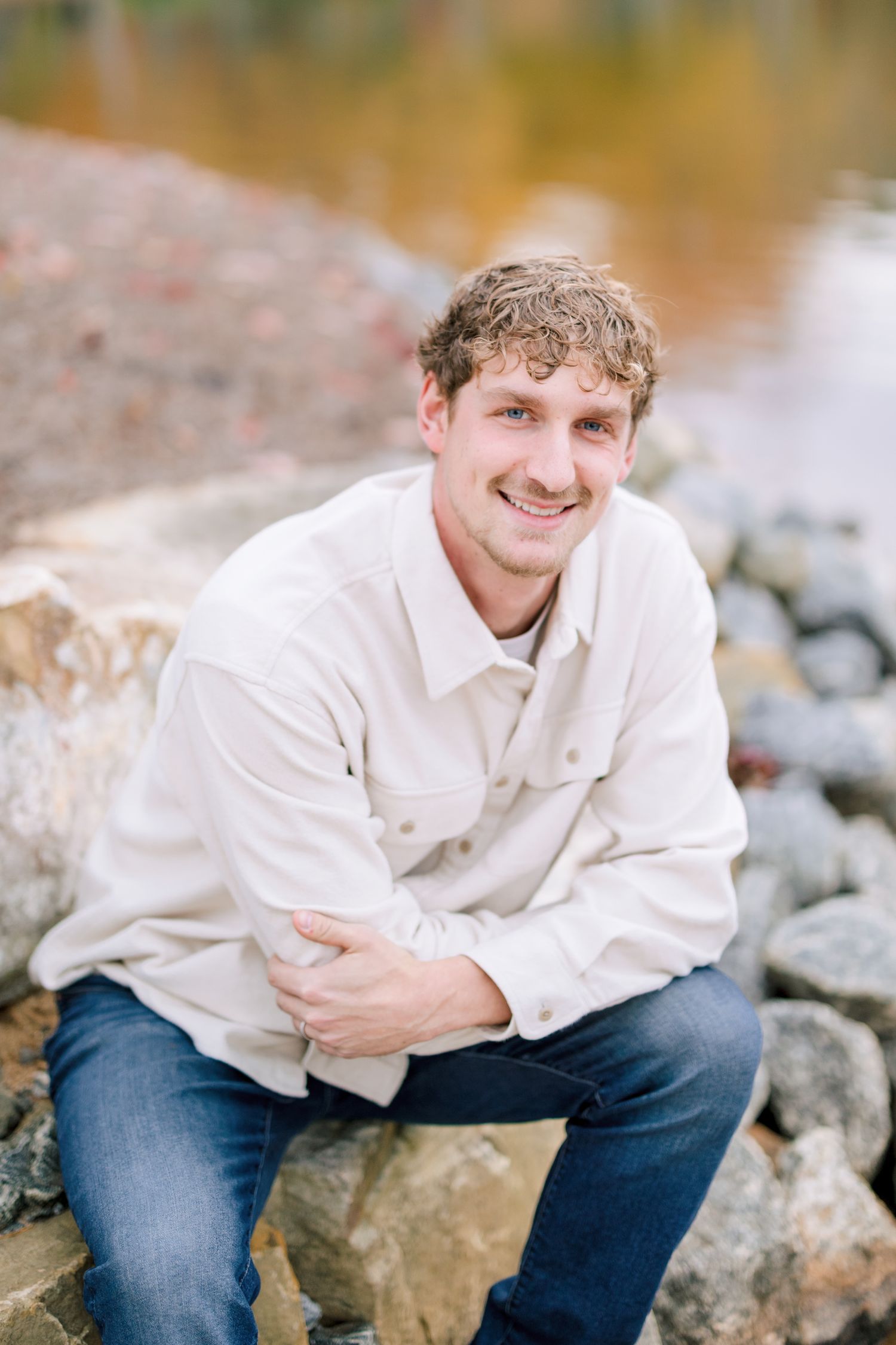 Portrait series on rocky shore wearing cream colored shirt against autumn background in Greensboro with a Family Photographer