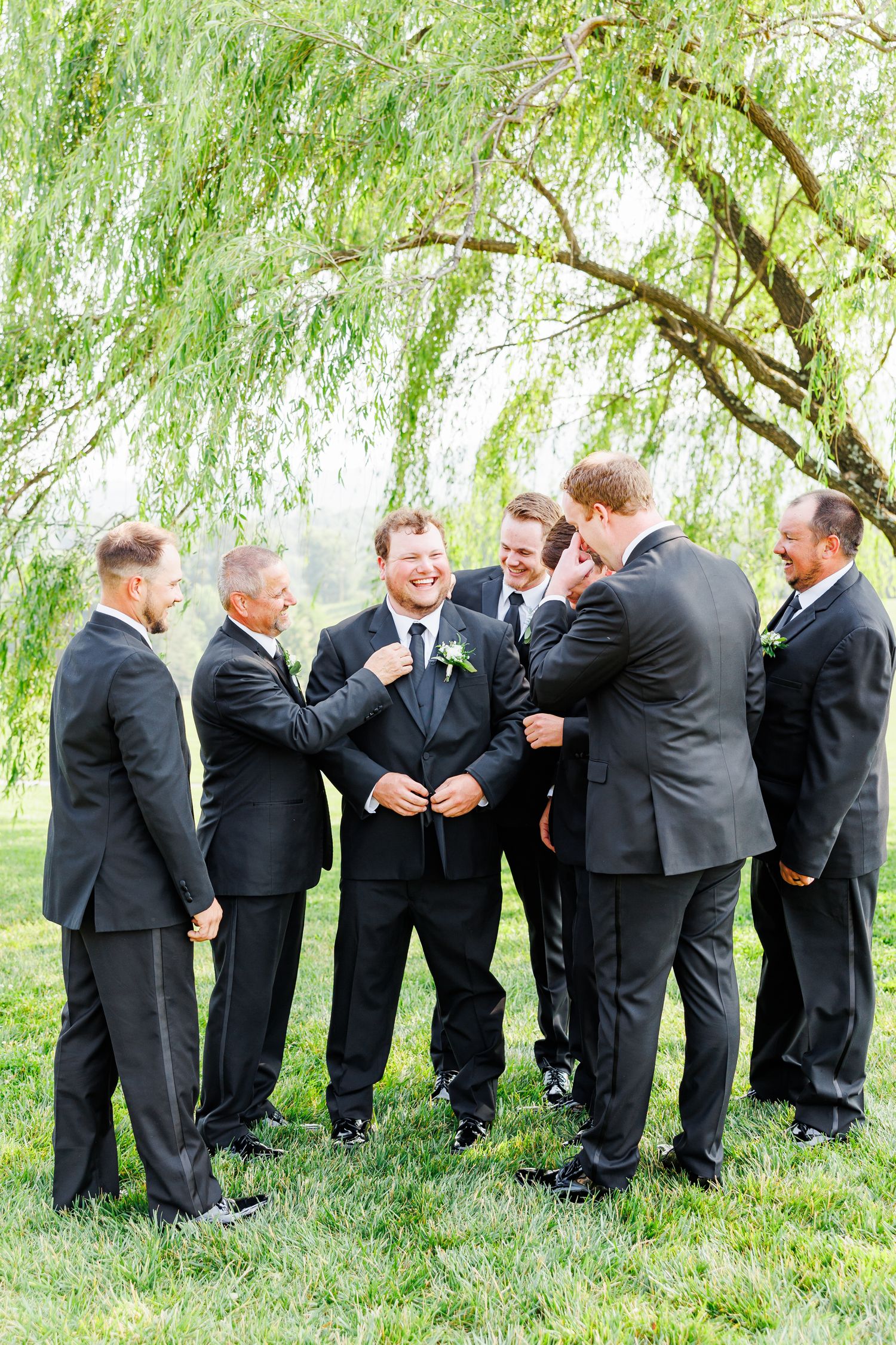 Groomsmen in black suits sharing laughs in a playful line under a willow tree at the Shilo with NC Wedding photographer