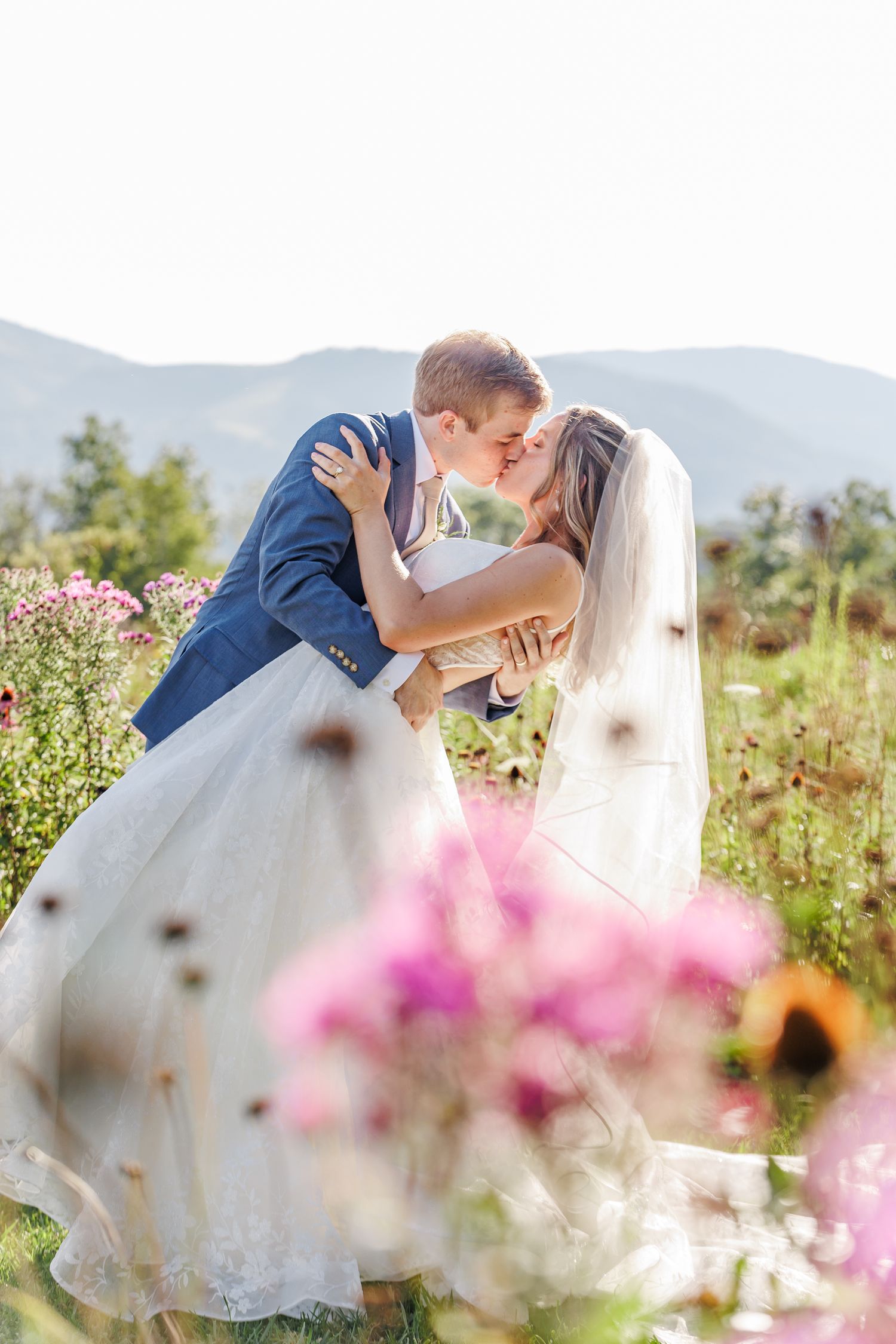 Romantic wedding couple embracing in field of wildflowers with mountains in background.