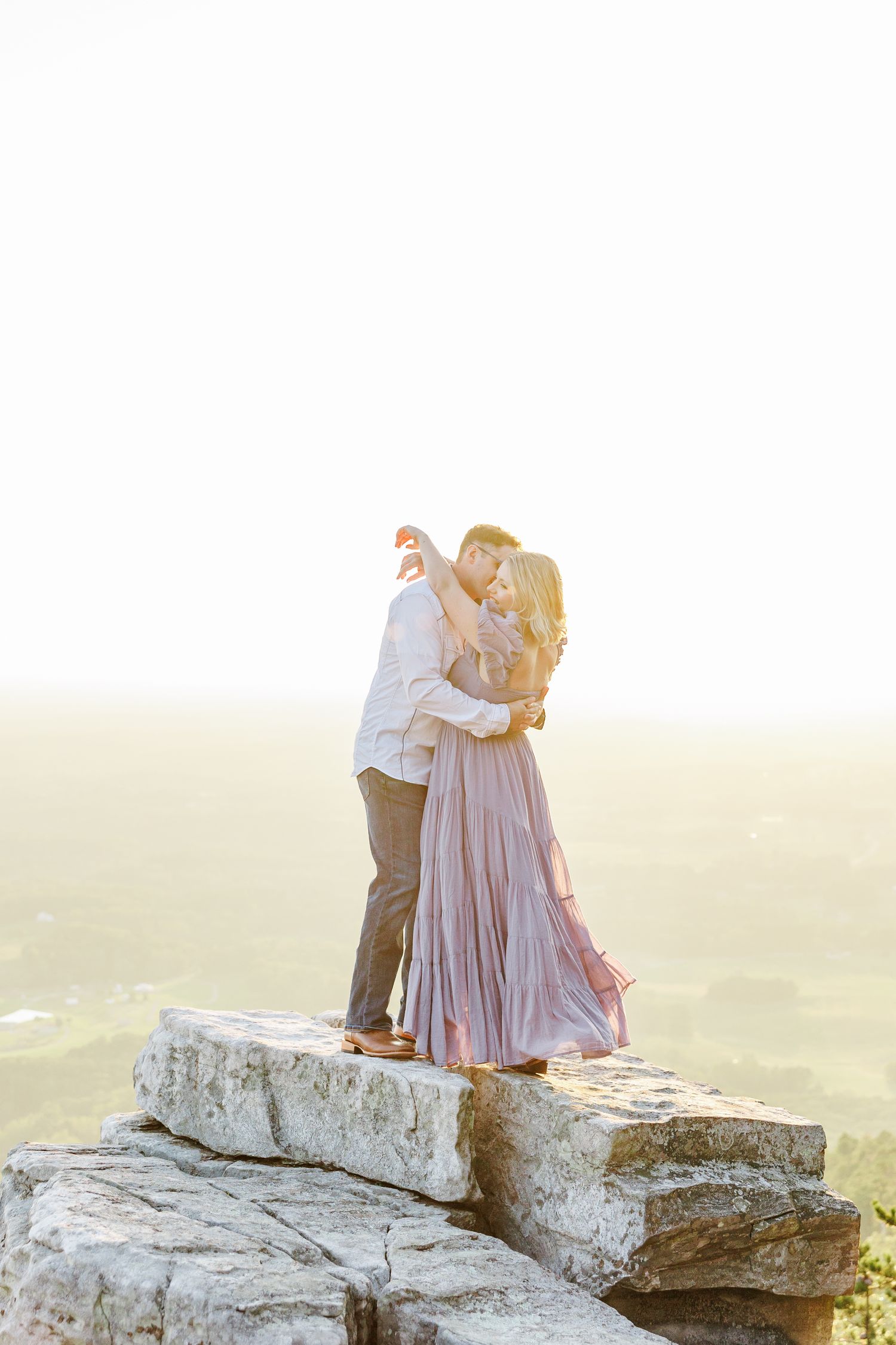 A couple embraces on a rocky cliff edge during golden hour with Winston Salem Engagement Photographer at Pilot Mountain