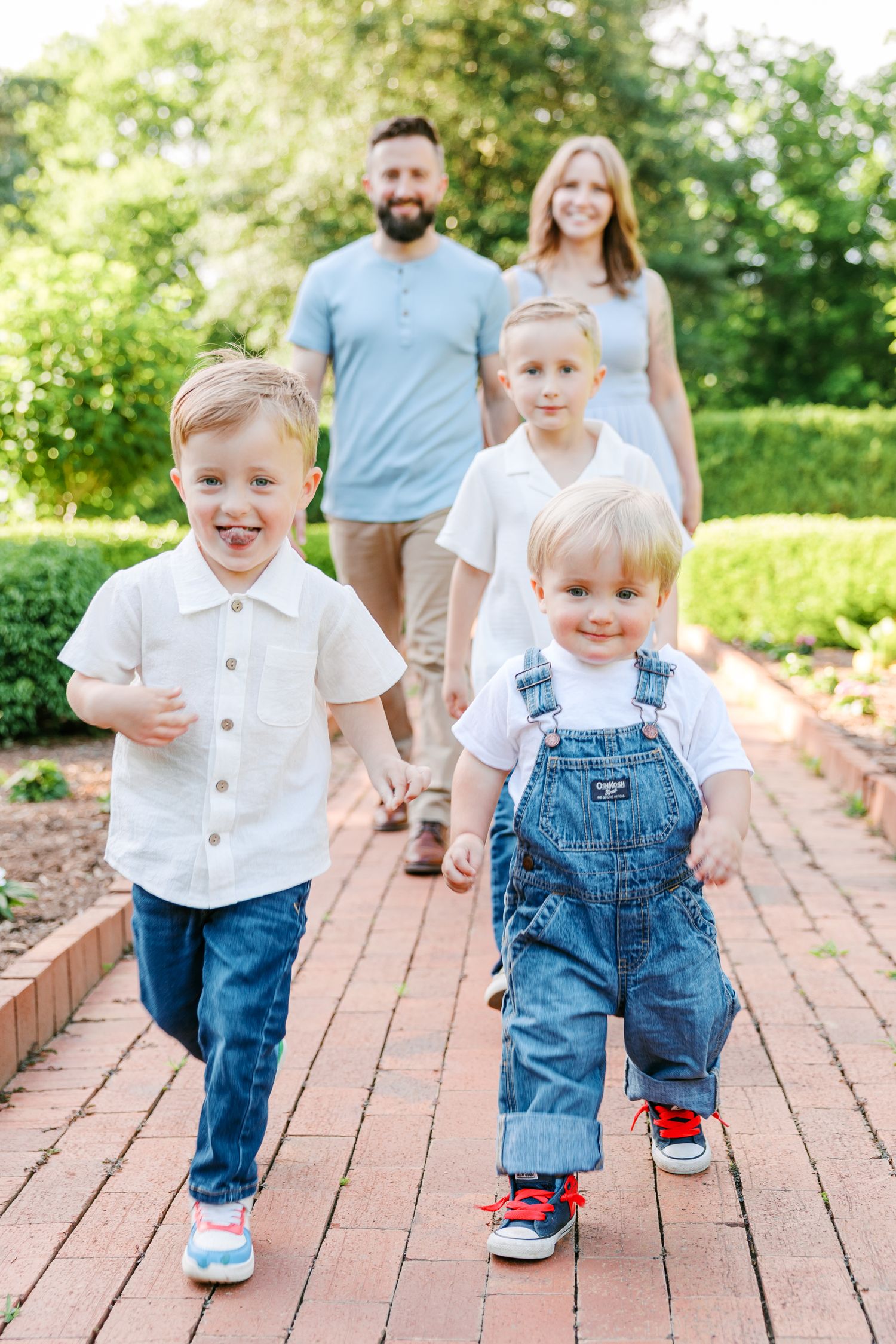 Toddler in overalls walking on brick path with family in background in Clemmons NC  with a Family Photographer