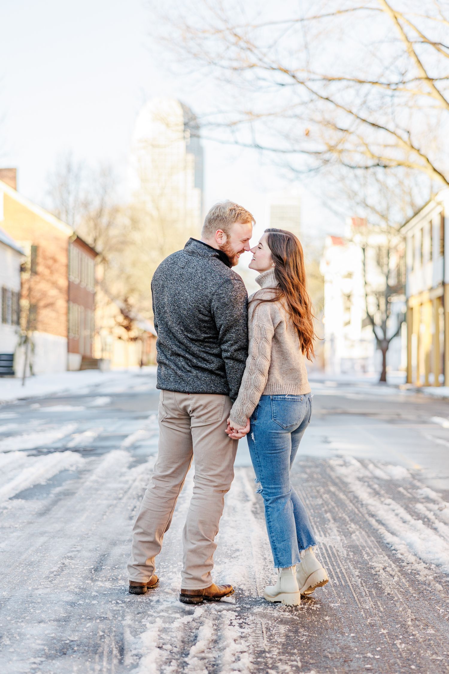 A couple shares an intimate moment on a snowy city street with buildings in the background with Winston Salem Engagement Photographer