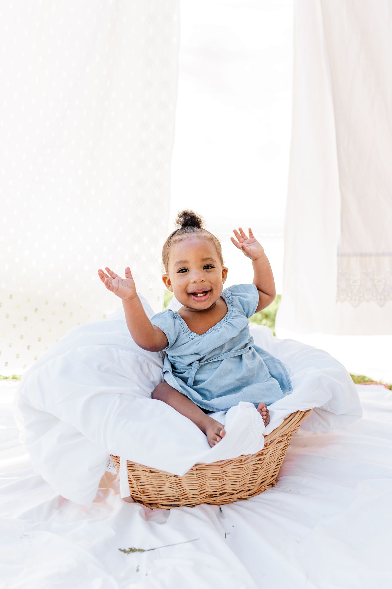 Happy baby in blue outfit playing in basket surrounded by white linens  in Winston Salem NC  with a Family Photographer