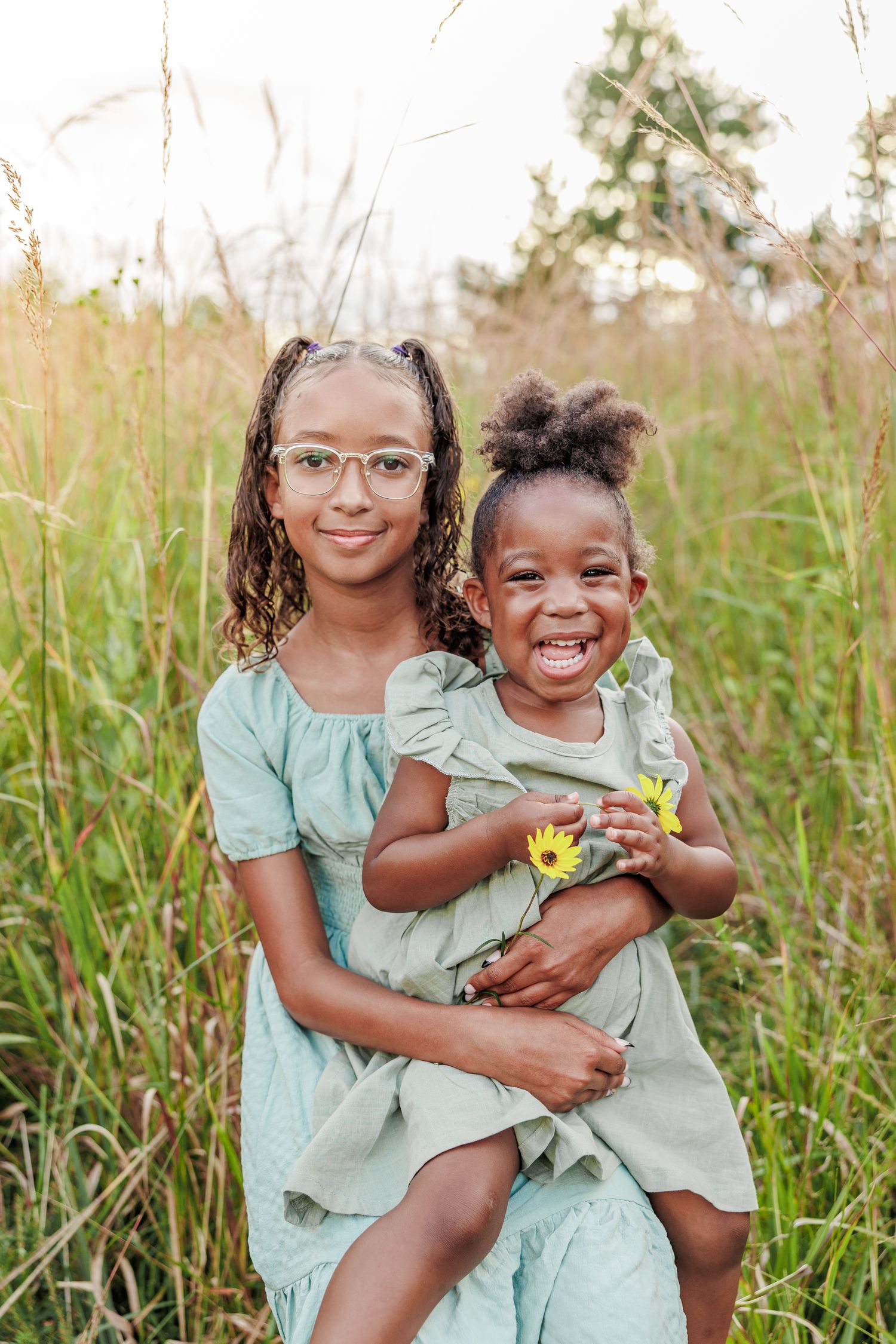 Two children in matching mint green dresses smile while playing in tall grass in Greensboro NC  with a Family Photographer.
