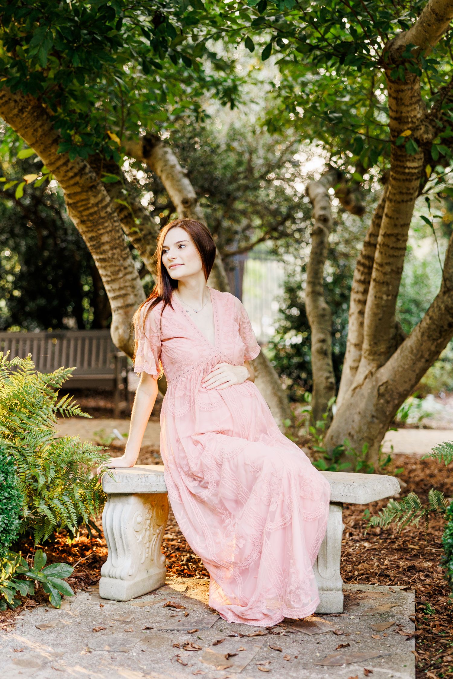 A woman in a pink lace dress sits gracefully on a stone bench under blooming trees in Clemmons NC  with a Maternity Photographer
