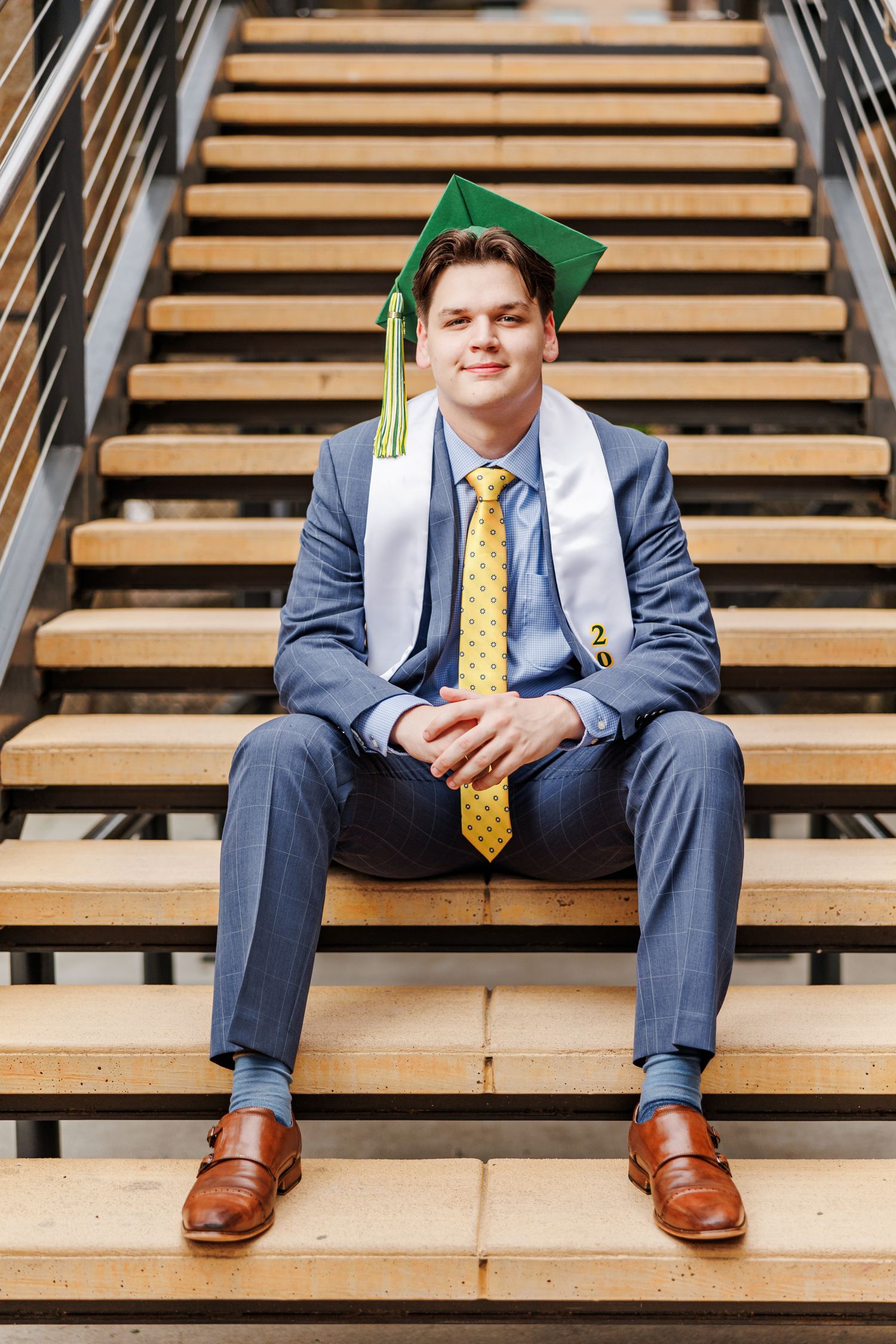 Graduate in blue suit and green cap sitting on wooden stairs for portrait  for Senior Portrait Session in Winston Salem