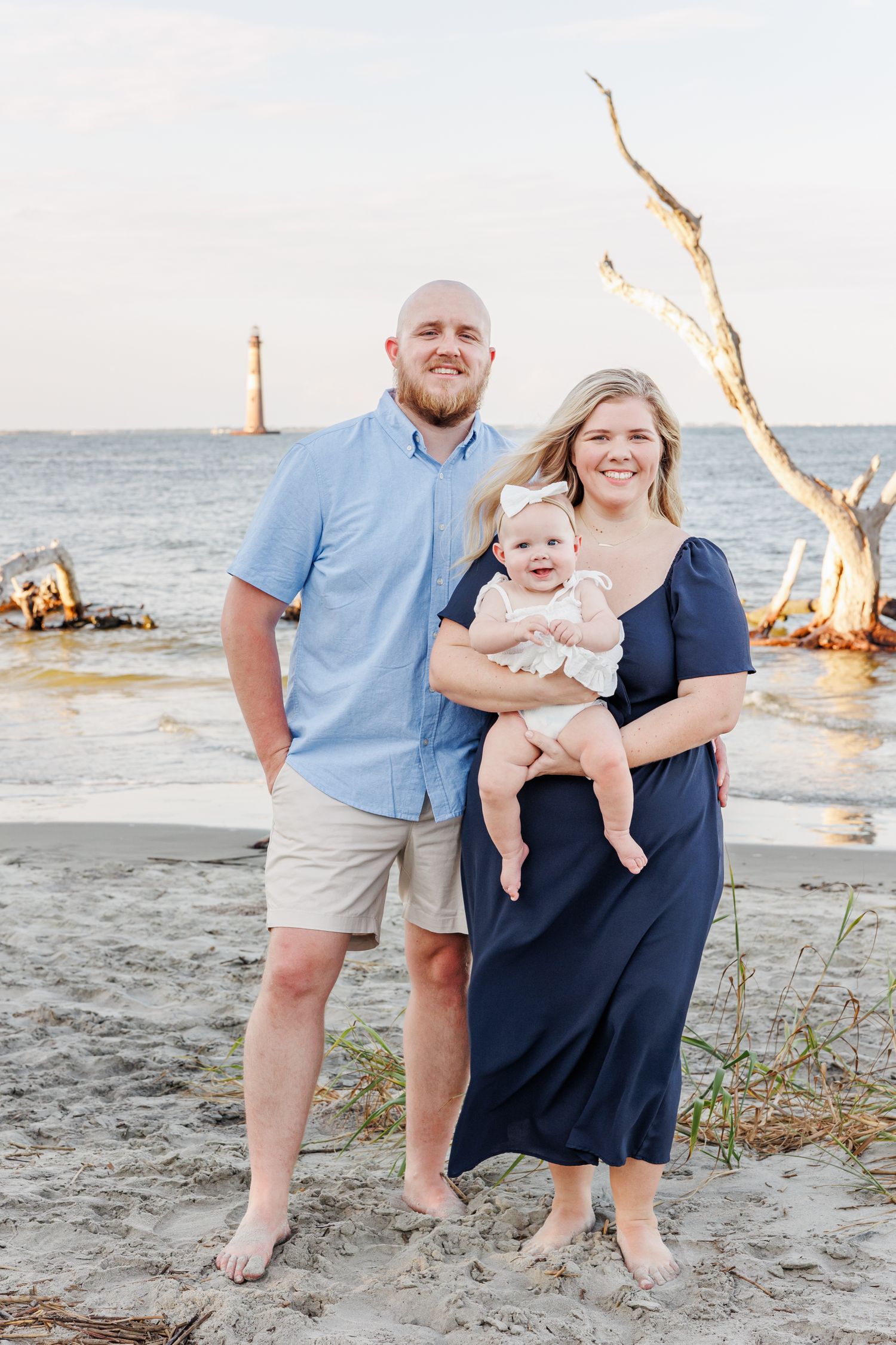 A family poses together on a beach at sunset with driftwood and lighthouse in background in Charleston with a SC Family Photographer