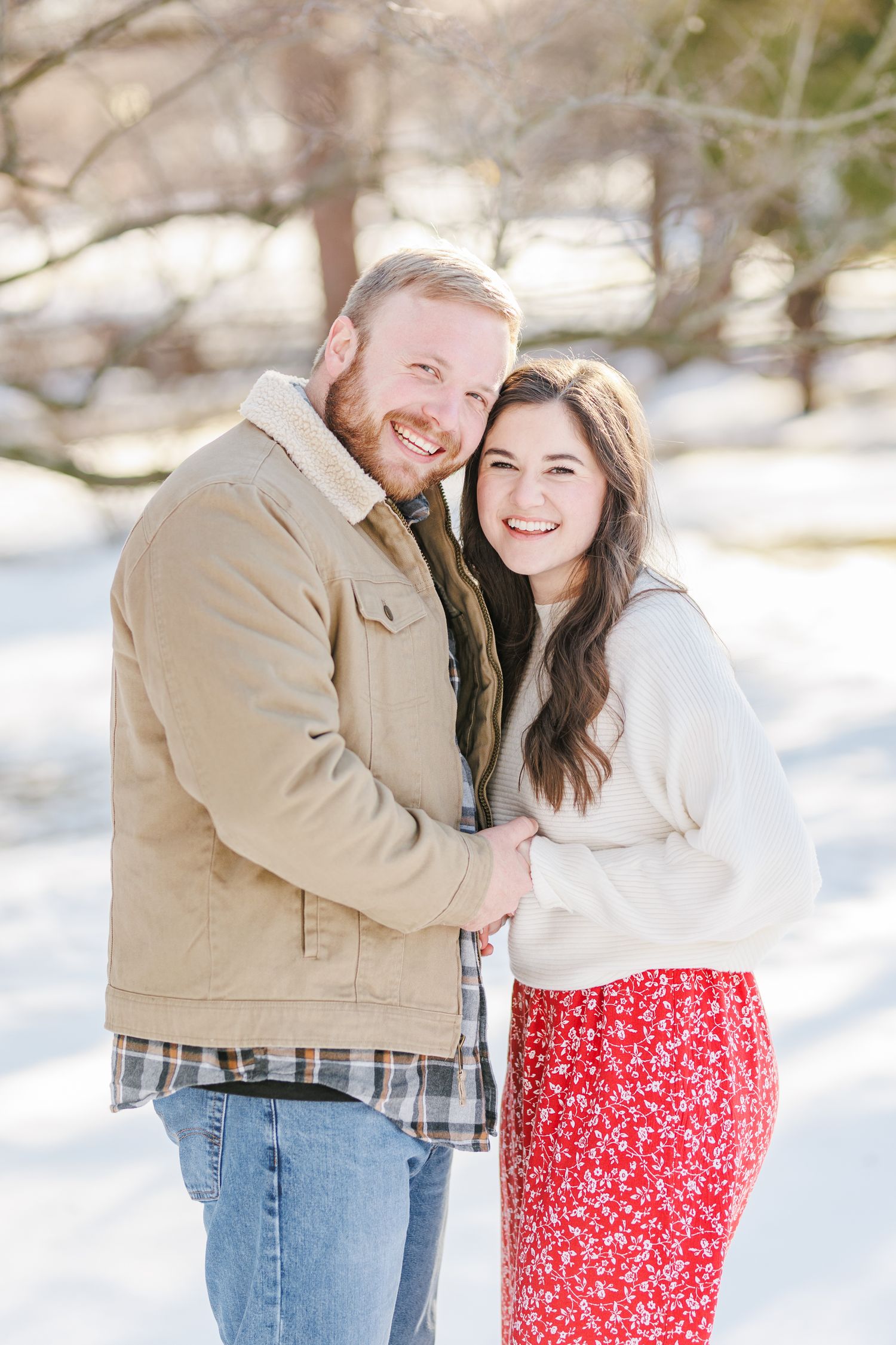 A couple embraces in a snowy winter setting wearing casual clothing and smiling at the camera with Winston Salem Engagement Photographer