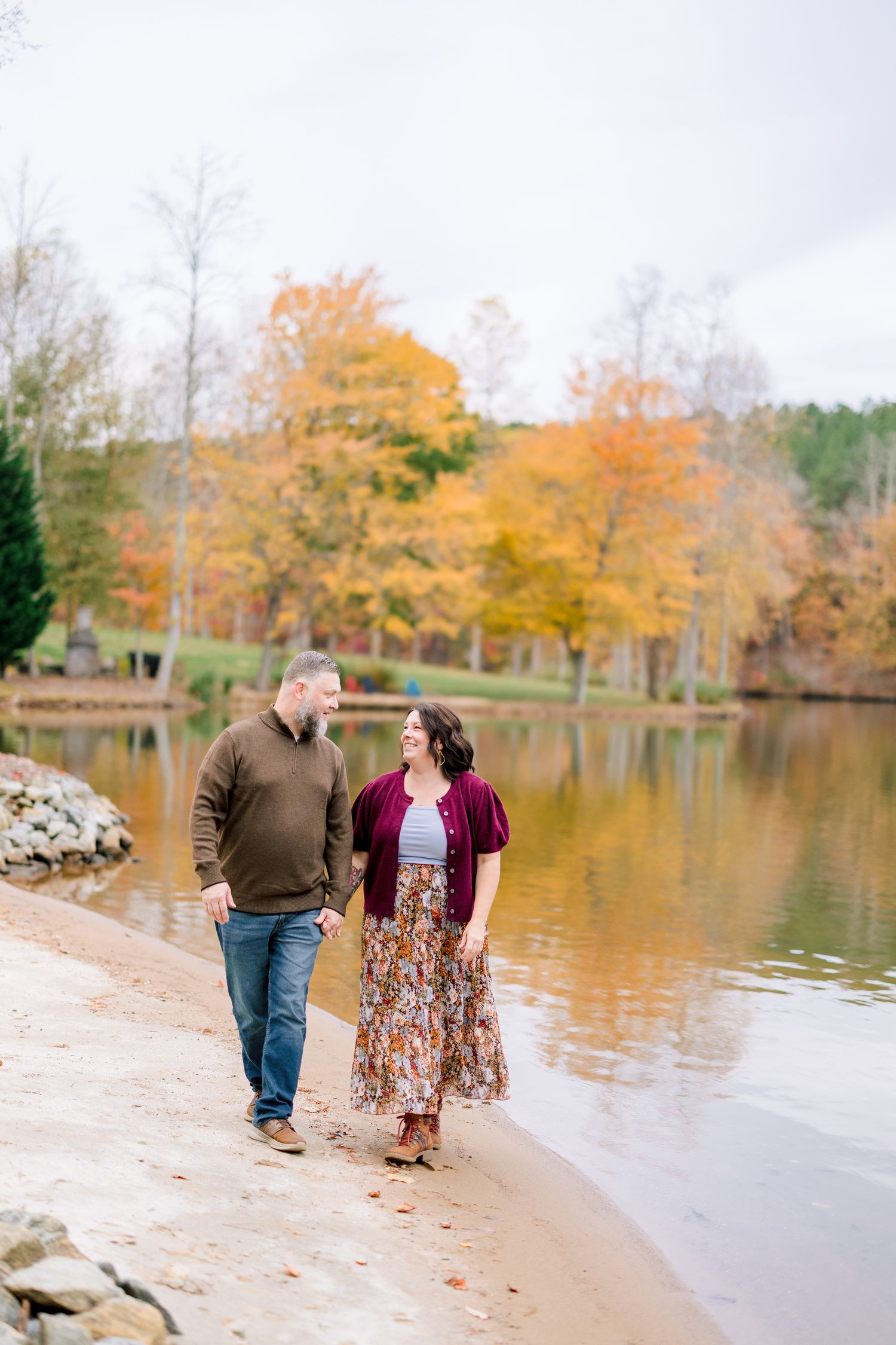 Couple walking along a scenic autumn lakeside path with golden foliage reflecting in the calm water in Greensboro with a Family Photographer