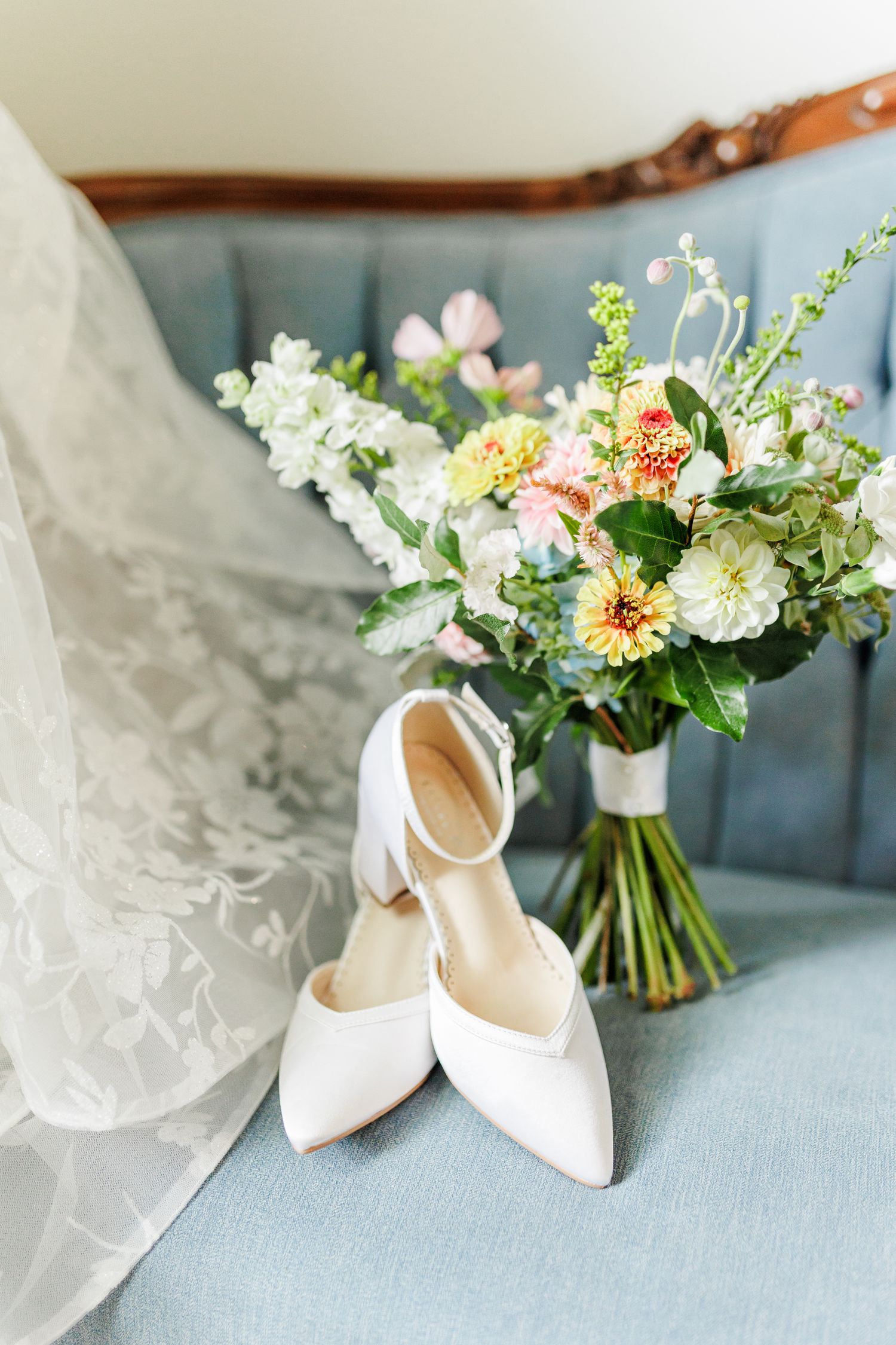 Elegant white bridal shoes and delicate floral bouquet arranged on light blue surface with flowing veil at Appalachian View Venue