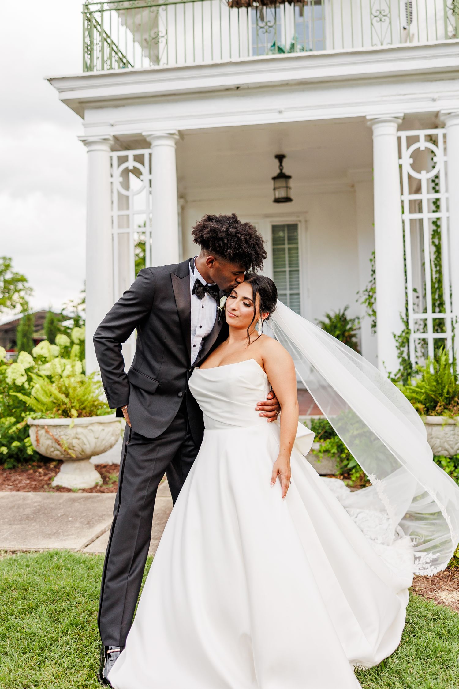 Romantic wedding couple poses against white pillars and greenery in an elegant outdoor setting at the Finch House in Thomasville