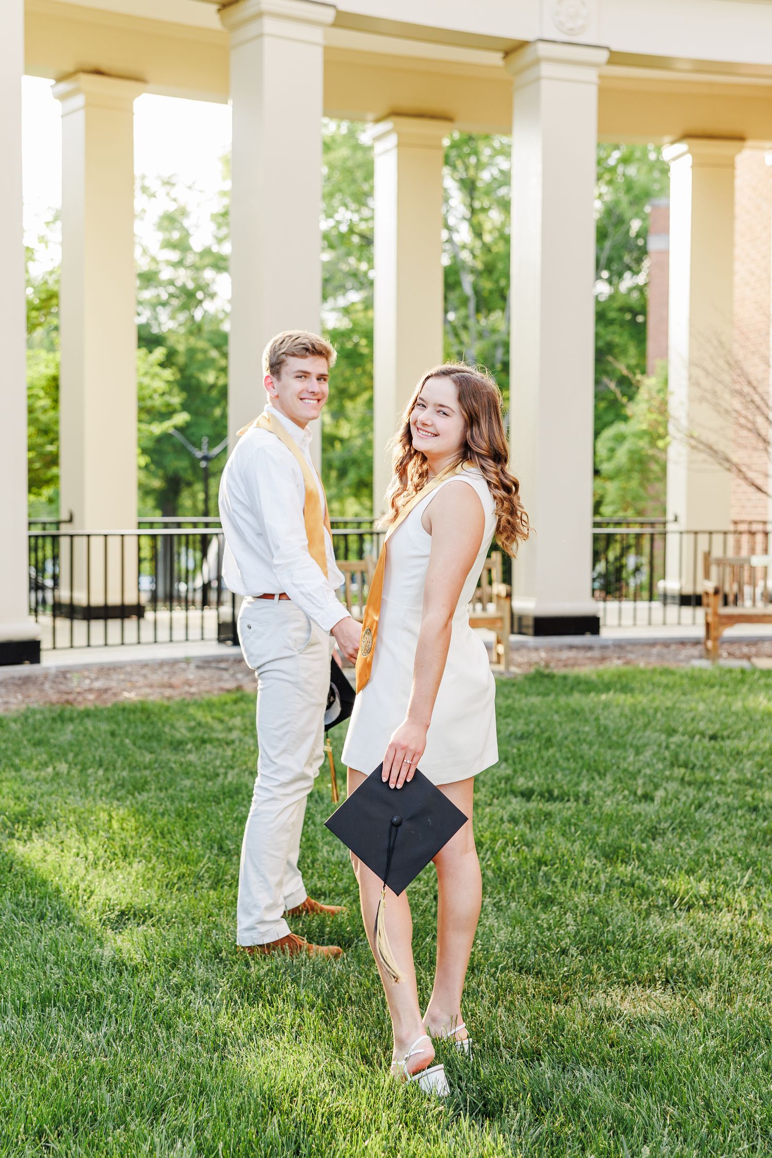 Couple in white clothing poses playfully on grass near columned building at Wake Forest University with a Graduation Photographer