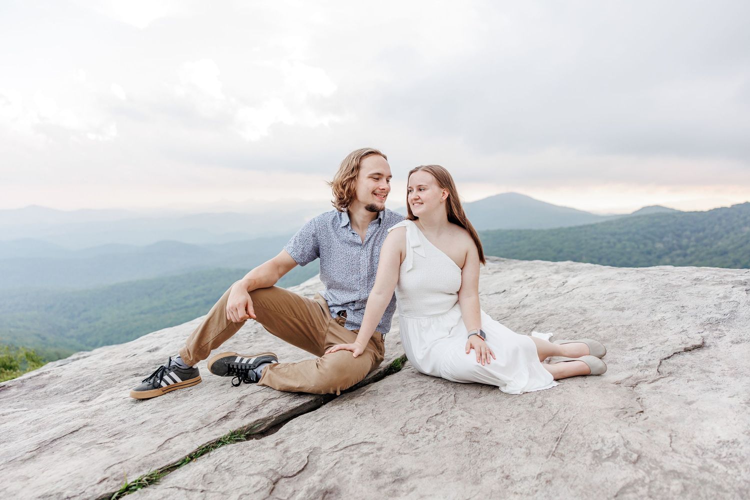 A couple sits together on a mountain summit with a scenic valley view with Blowing Rock Engagement Photographer on the Blue Ridge Parkway