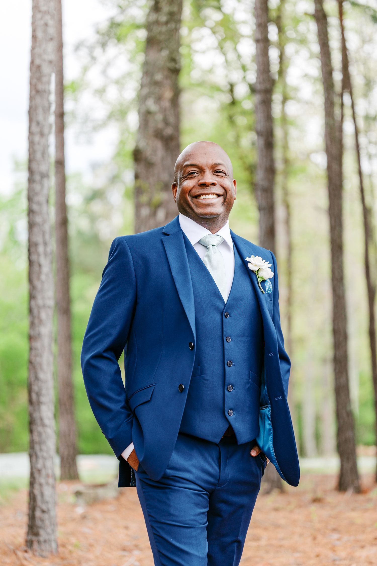 A groom in a navy blue three-piece suit smiles while standing in a wooded outdoor setting at the Evermore with Cary NC Wedding Photographer