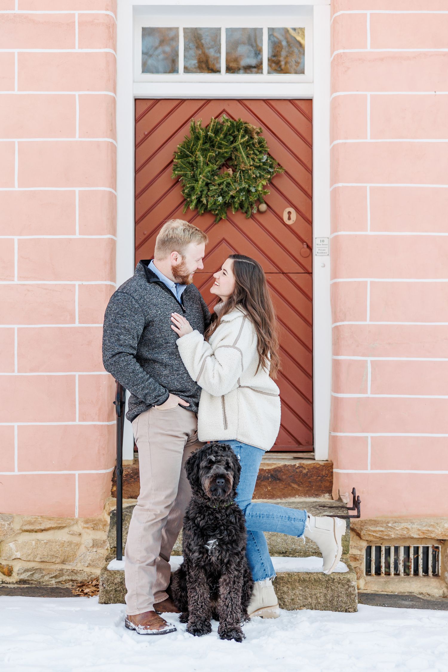 A couple poses with their black dog in front of a pink building with a holiday wreath with Winston Salem Engagement Photographer