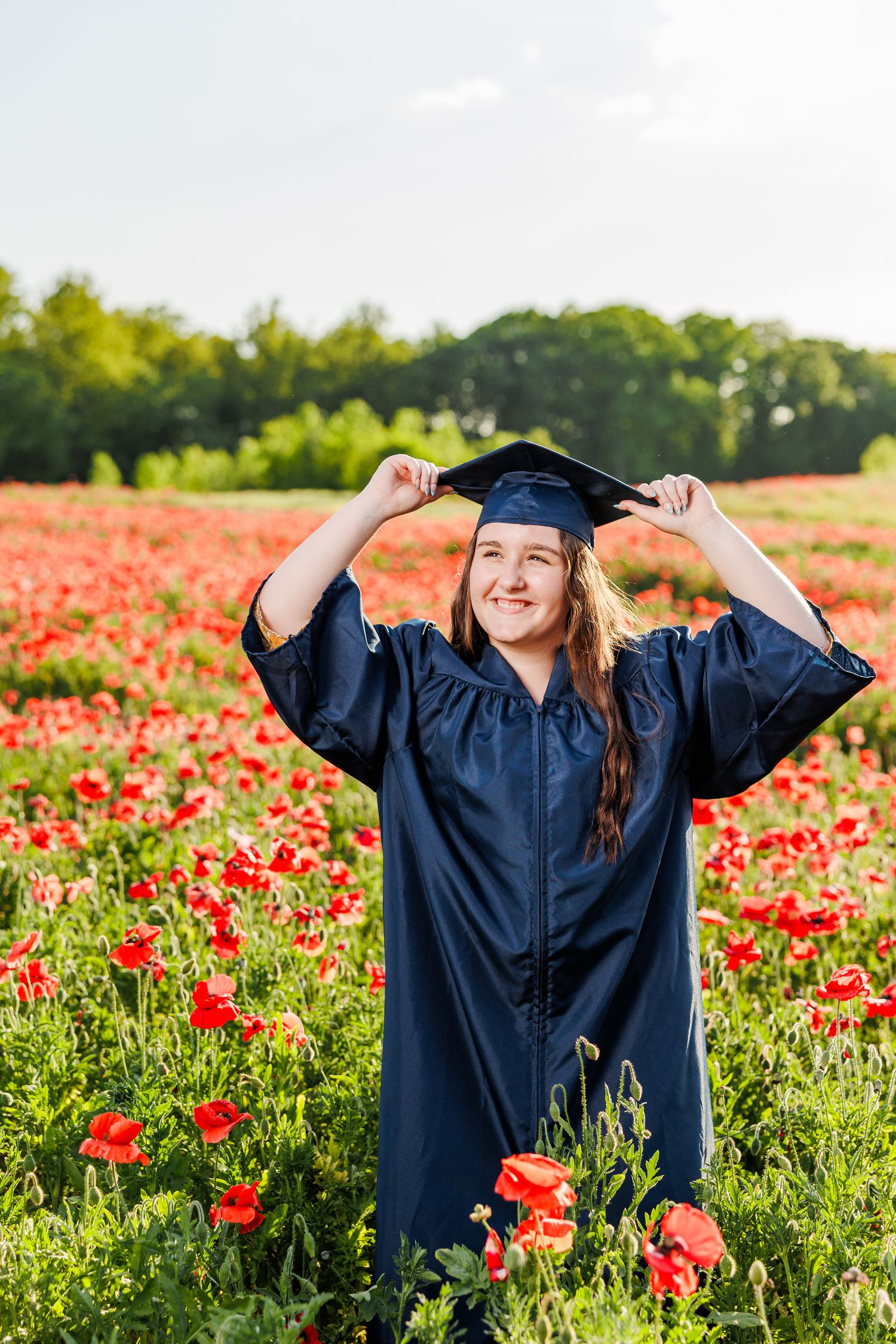 Graduate in navy blue cap and gown poses joyfully in a vibrant red poppy field during sunset  for Senior Portrait Session in Winston Salem