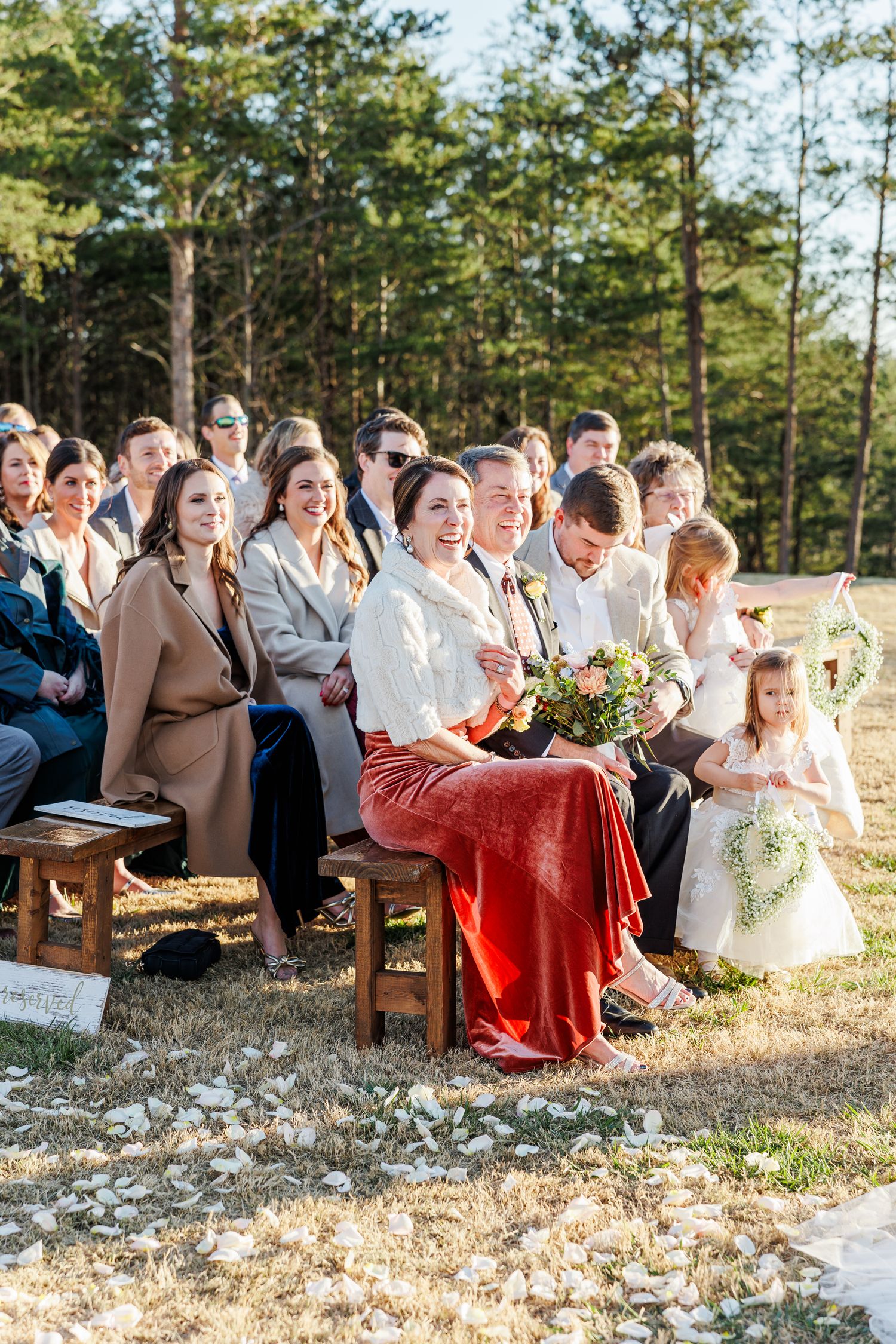 Wedding guests seated on wooden benches for an outdoor ceremony among pine trees.