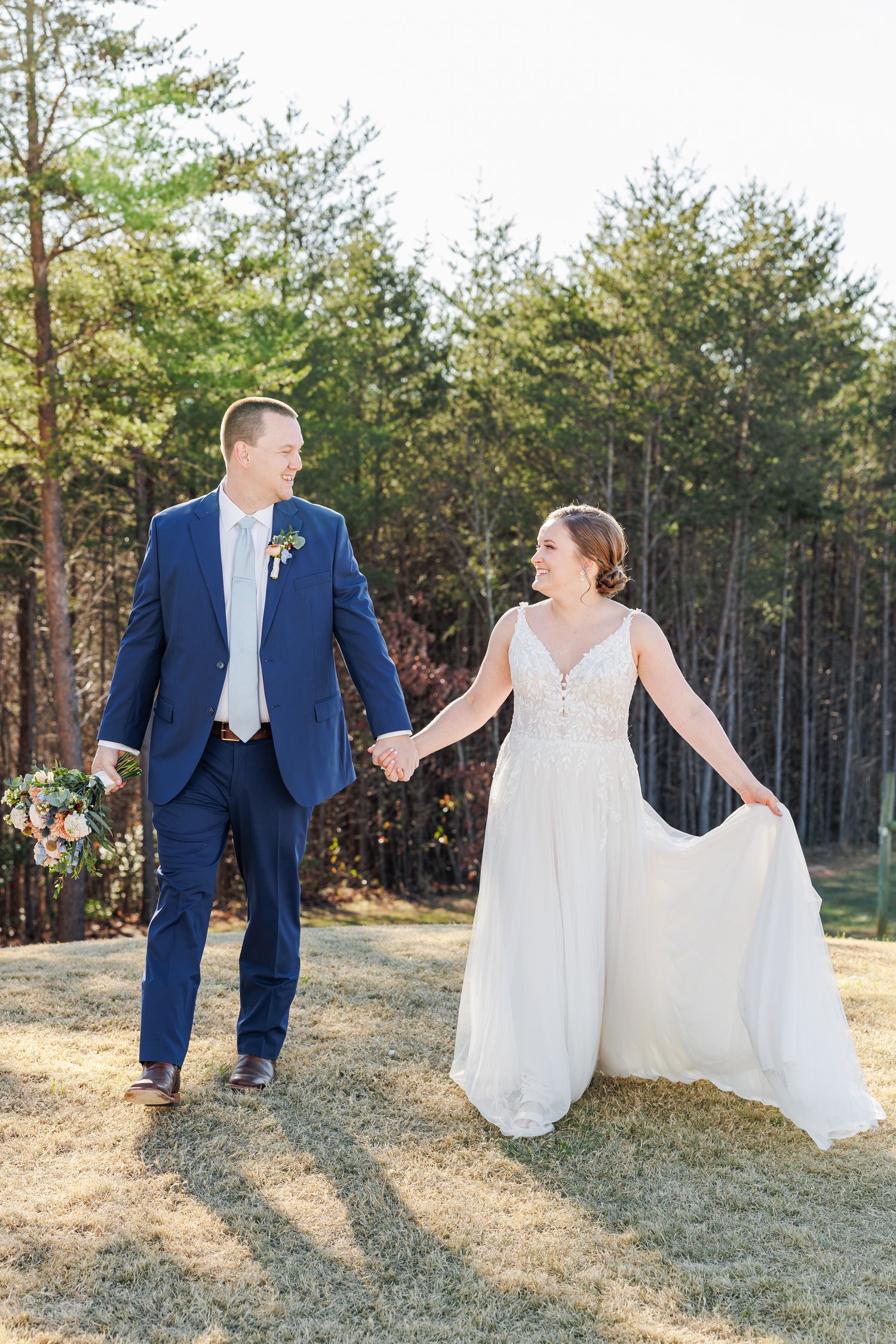 A couple in wedding attire poses playfully in a grassy field with pine trees in the background.