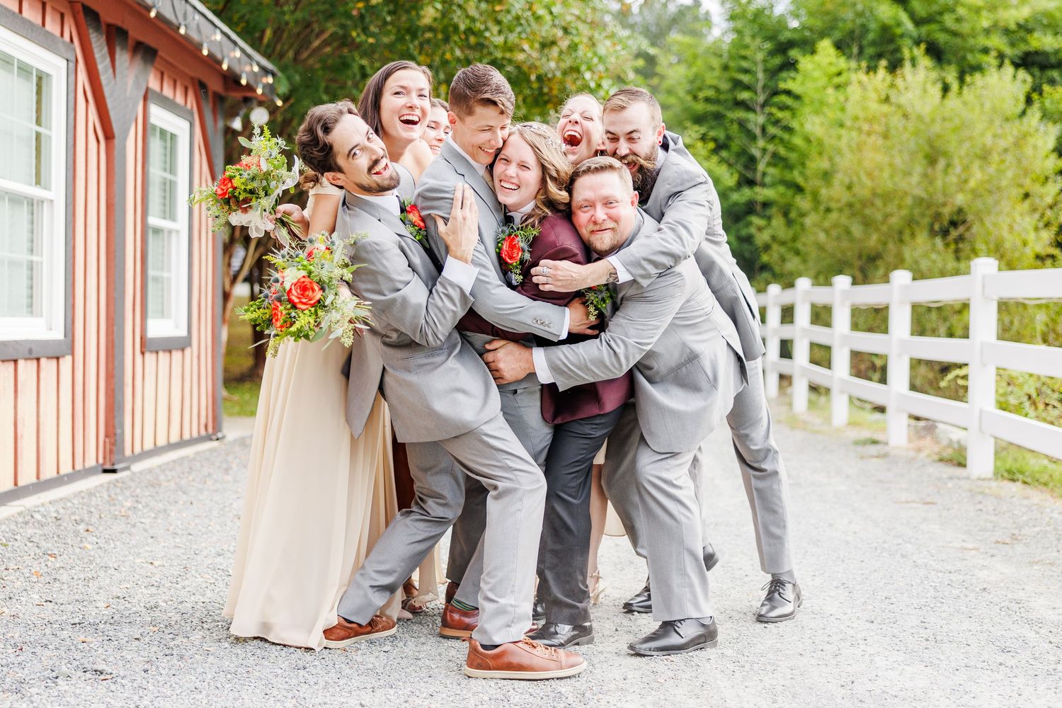 Wedding party poses for fun group photo on gravel path with white fence with NC Wedding Photographer