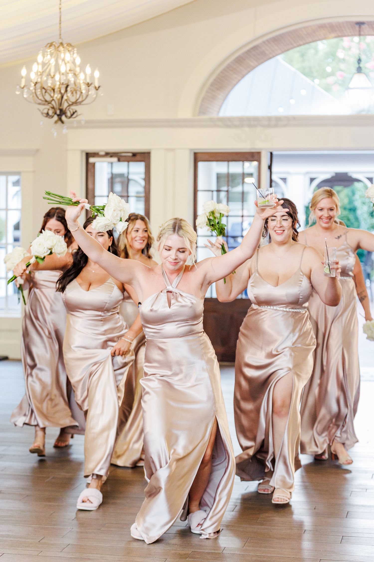 Bridesmaids in champagne satin dresses dance and laugh together in an elegant reception hall with Chapel Hill Wedding Photographer