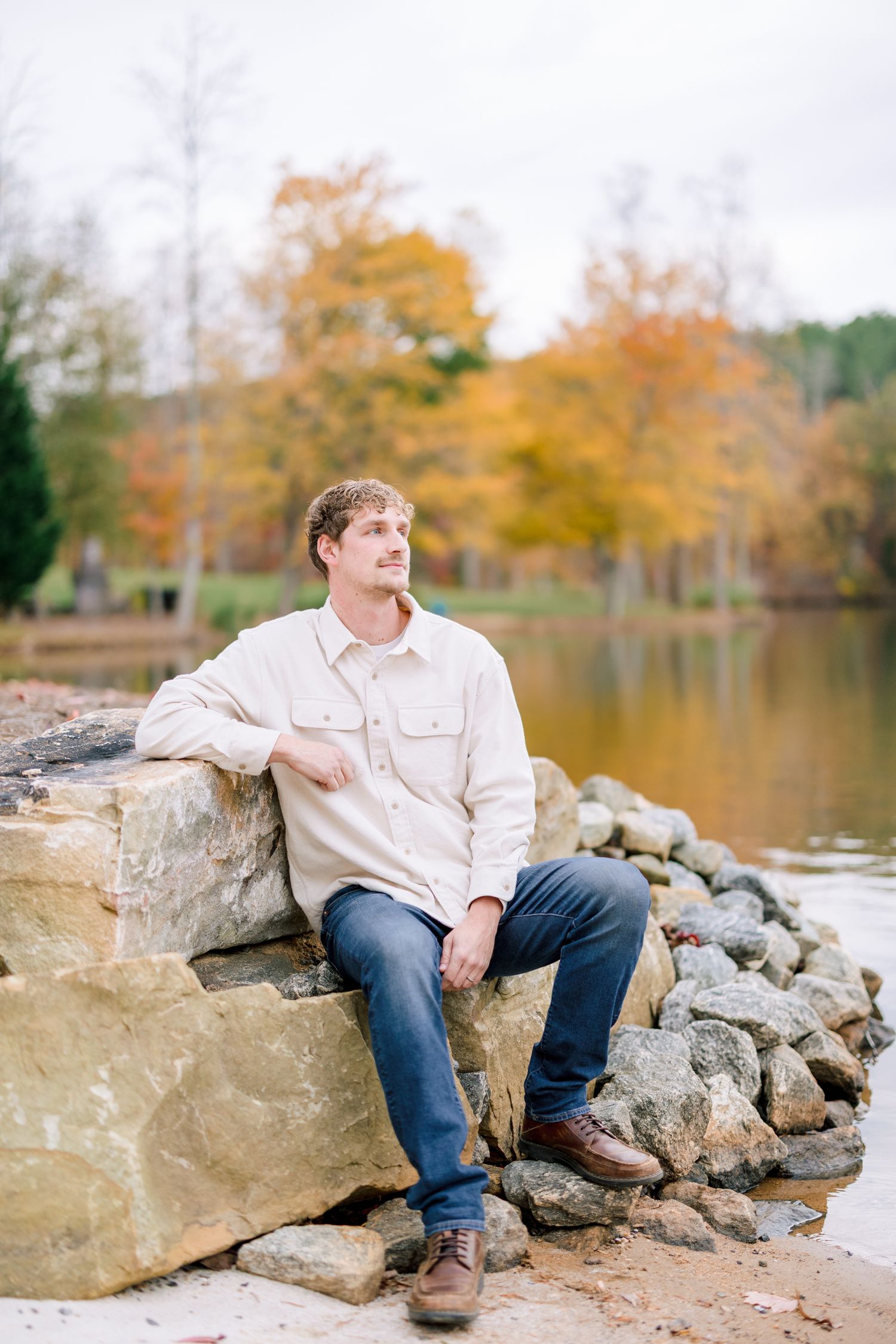 Casual portraits on rocky lakeside wearing light colored shirt with fall colors in background in Greensboro with a Family Photographer
