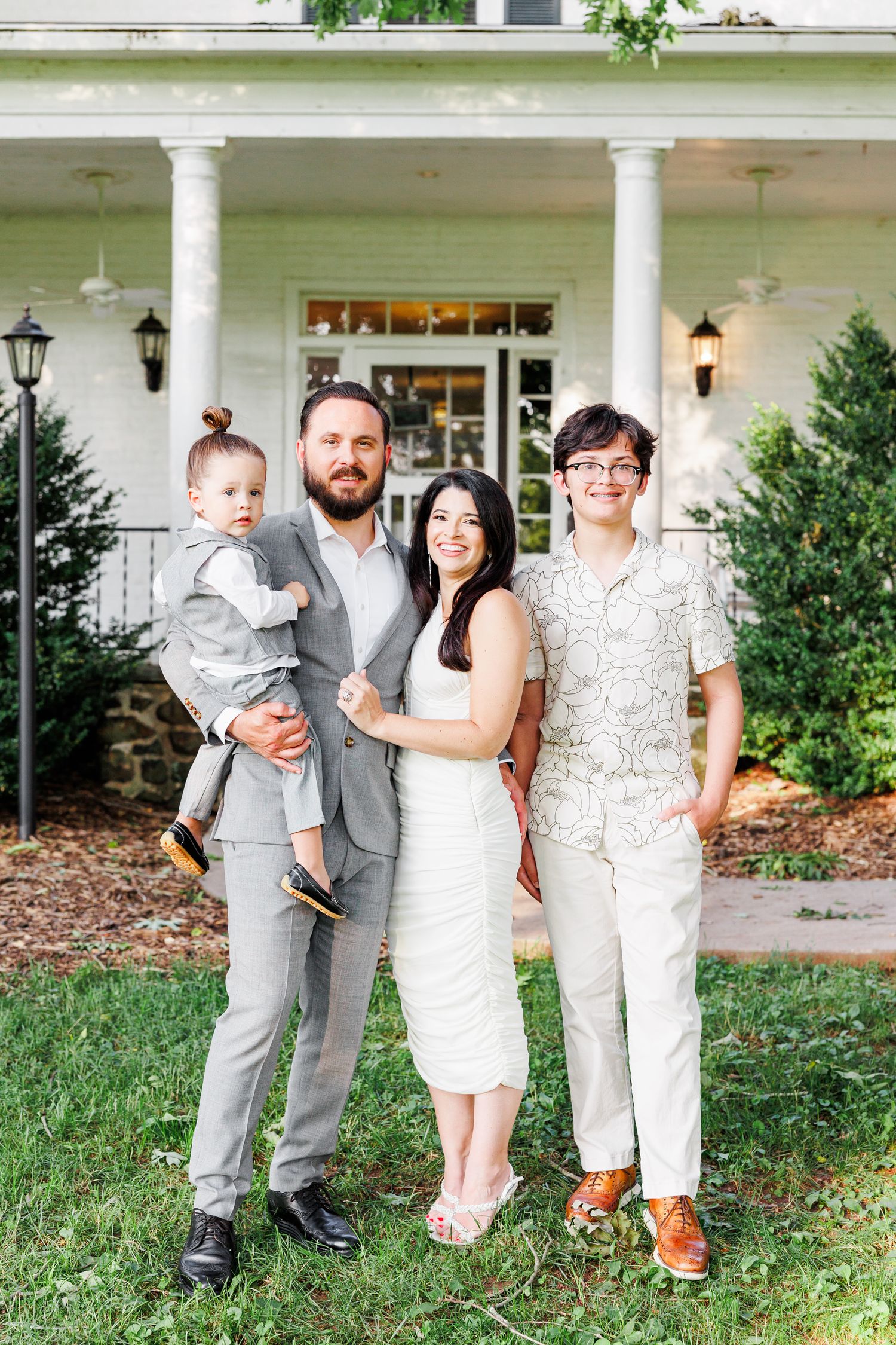 Family group poses together in light colored outfits on the lawn of a white columned porch with family portrait photographer in NC