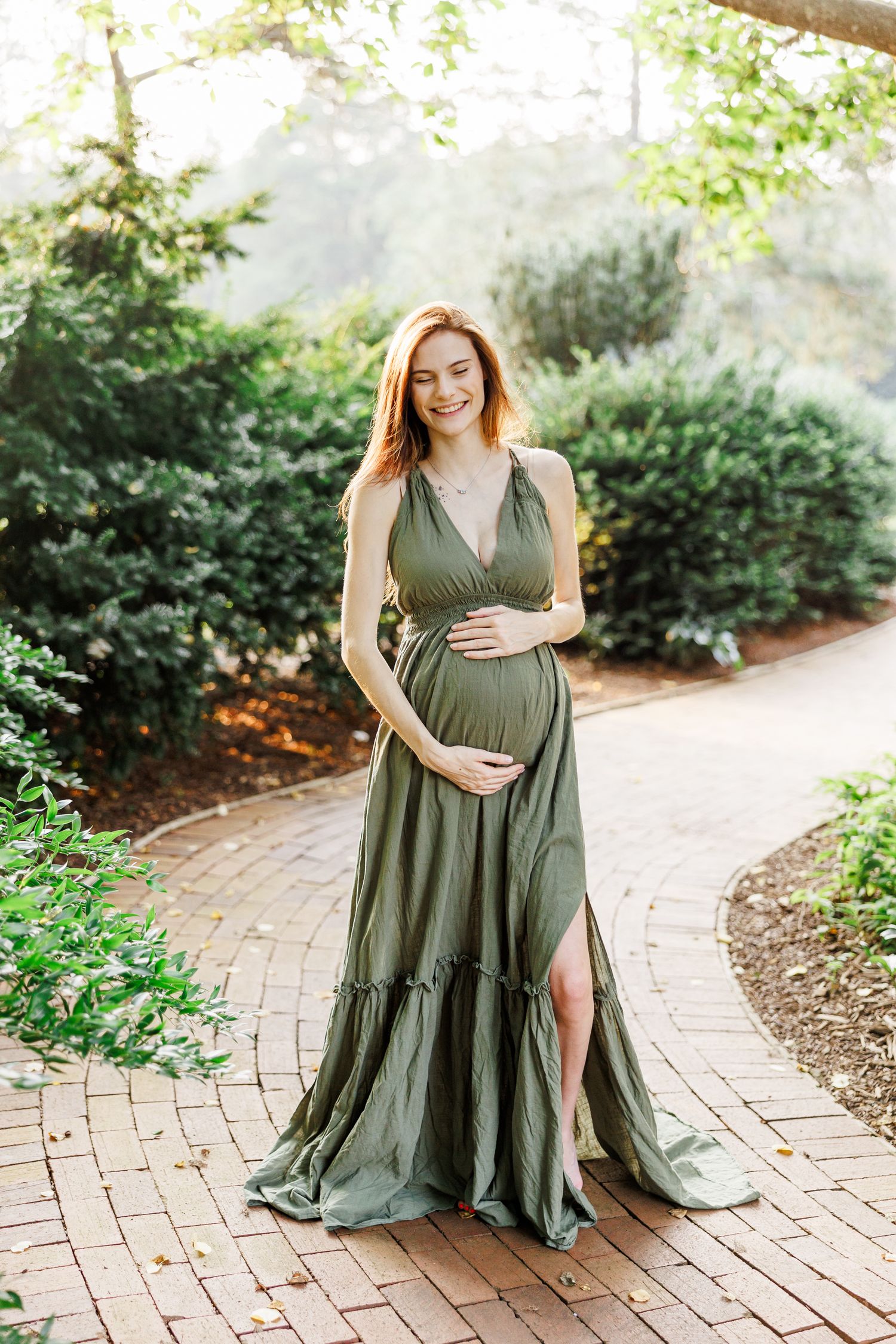 A woman in an olive green maxi dress poses on a brick path surrounded by greenery in Clemmons NC  with a Maternity Photographer